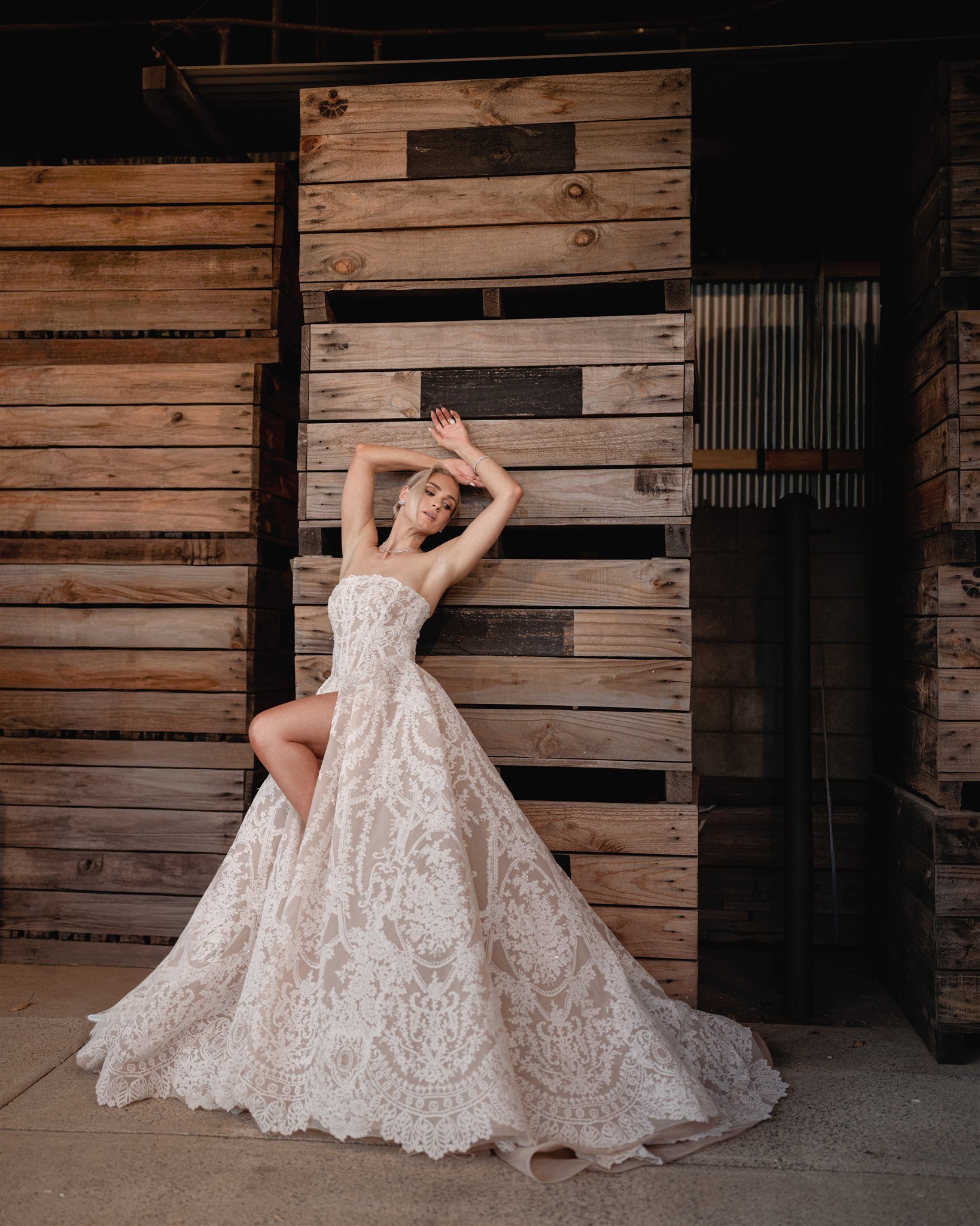 A woman in a white lace wedding dress poses against a wooden wall at night.