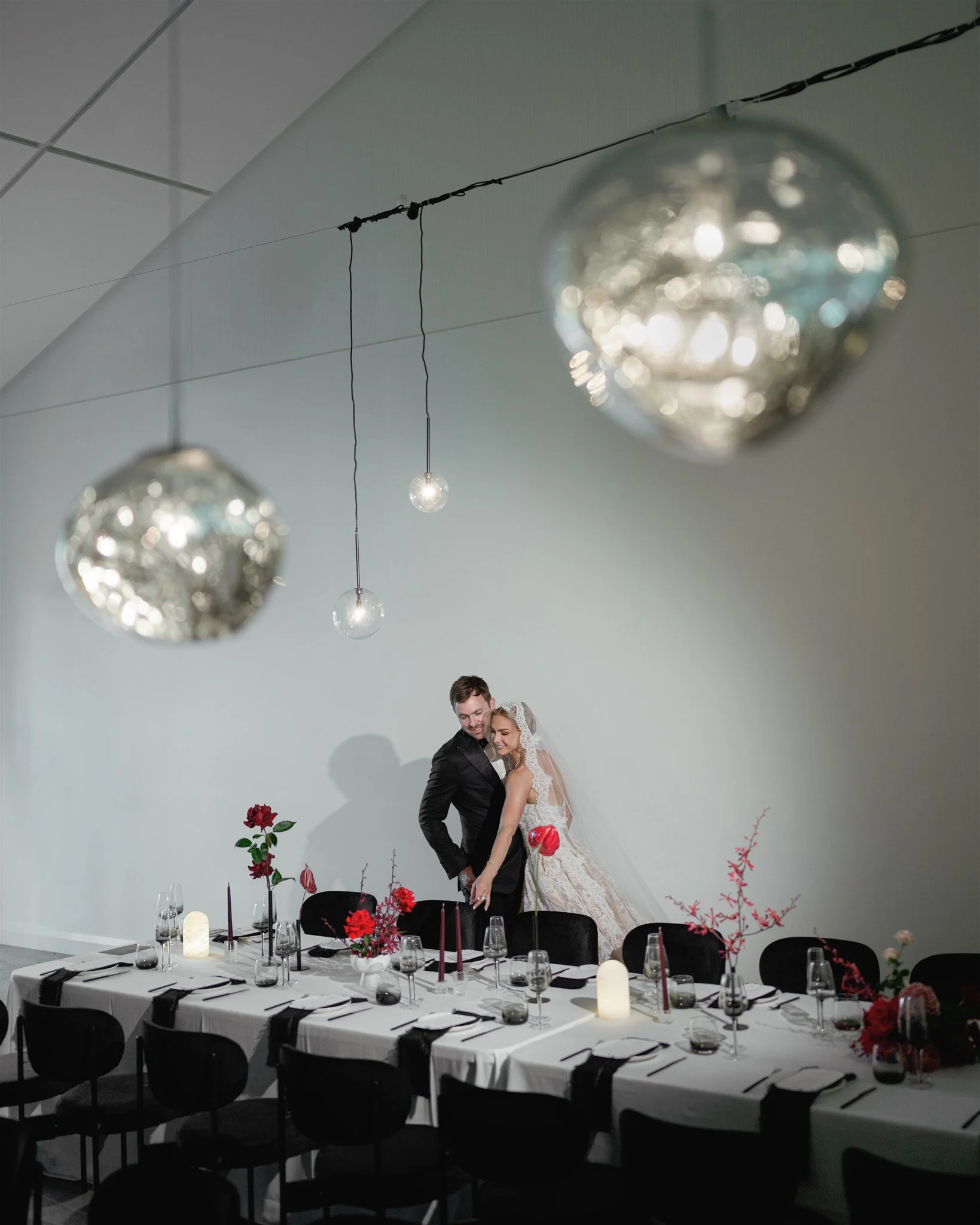 A bride and groom stand close together at a decorated wedding reception table, with the bride wearing a wedding dress and veil, and the groom in a black tuxedo, in a minimalist decorated indoor space with hanging light fixtures.