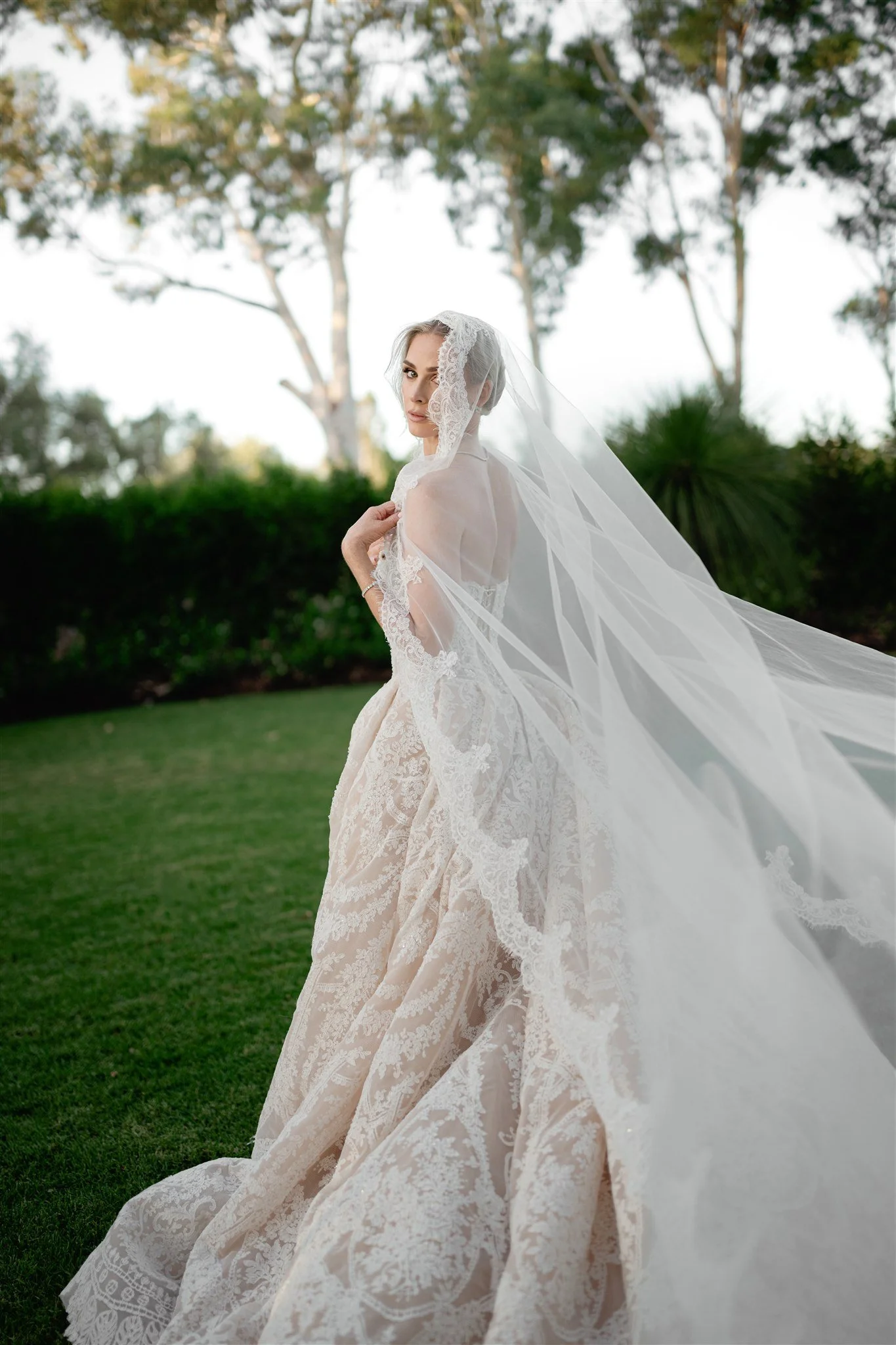 A bride in a lace wedding gown and veil standing outdoors on a grassy area with trees in the background.