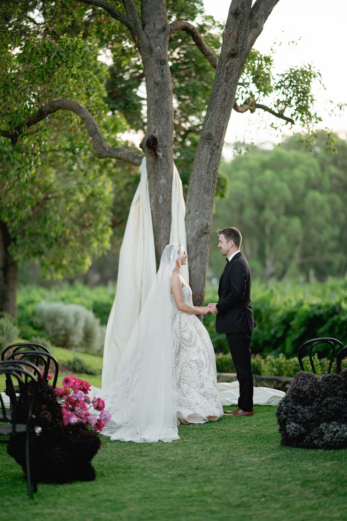 A bride and groom holding hands, standing outdoors during their wedding ceremony, under a large tree with draped fabric, surrounded by greenery and chairs.