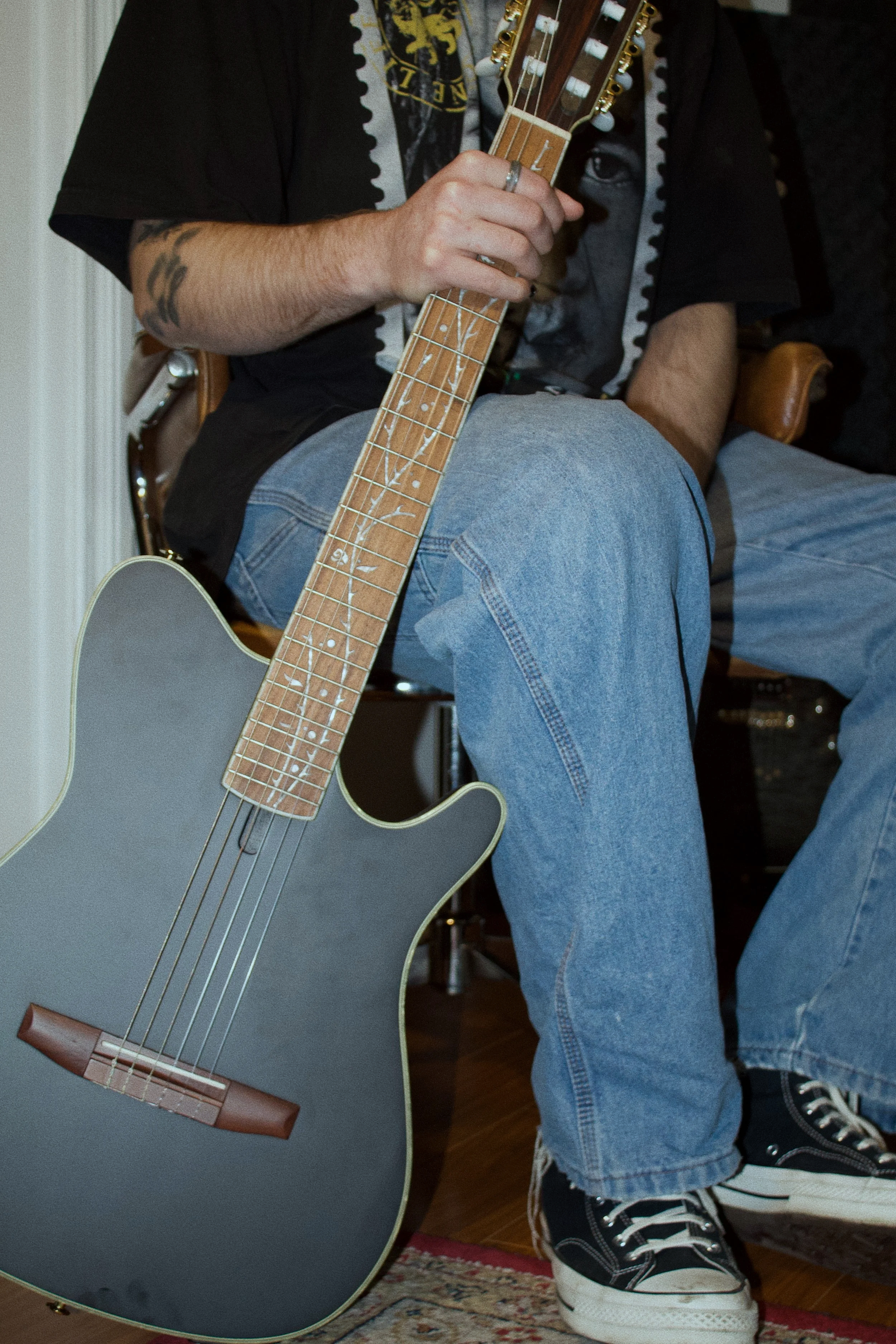 A person sitting on a chair, holding the neck of an acoustic guitar with a black body and a wooden fretboard. They are wearing a black t-shirt with a graphic print, light blue jeans, and black and white sneakers, with a tattoo on their forearm.