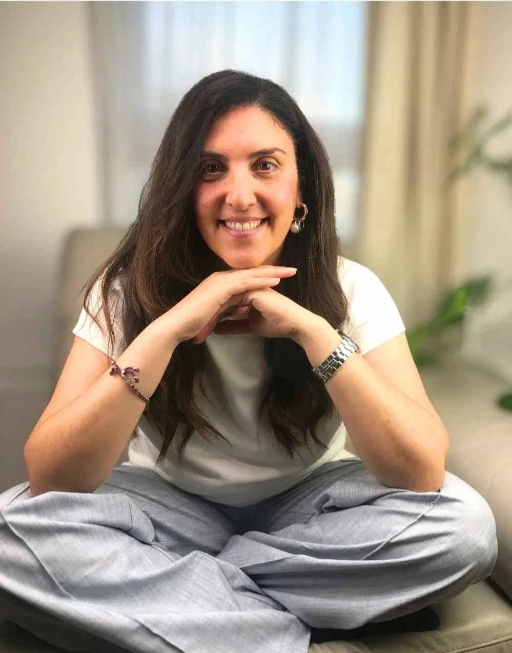 Una donna con capelli castani e lunghi, sorridente, seduta con le gambe incrociate su un divano, con mani appoggiate sotto il mento, in un ambiente luminoso con tende e piante sullo sfondo.