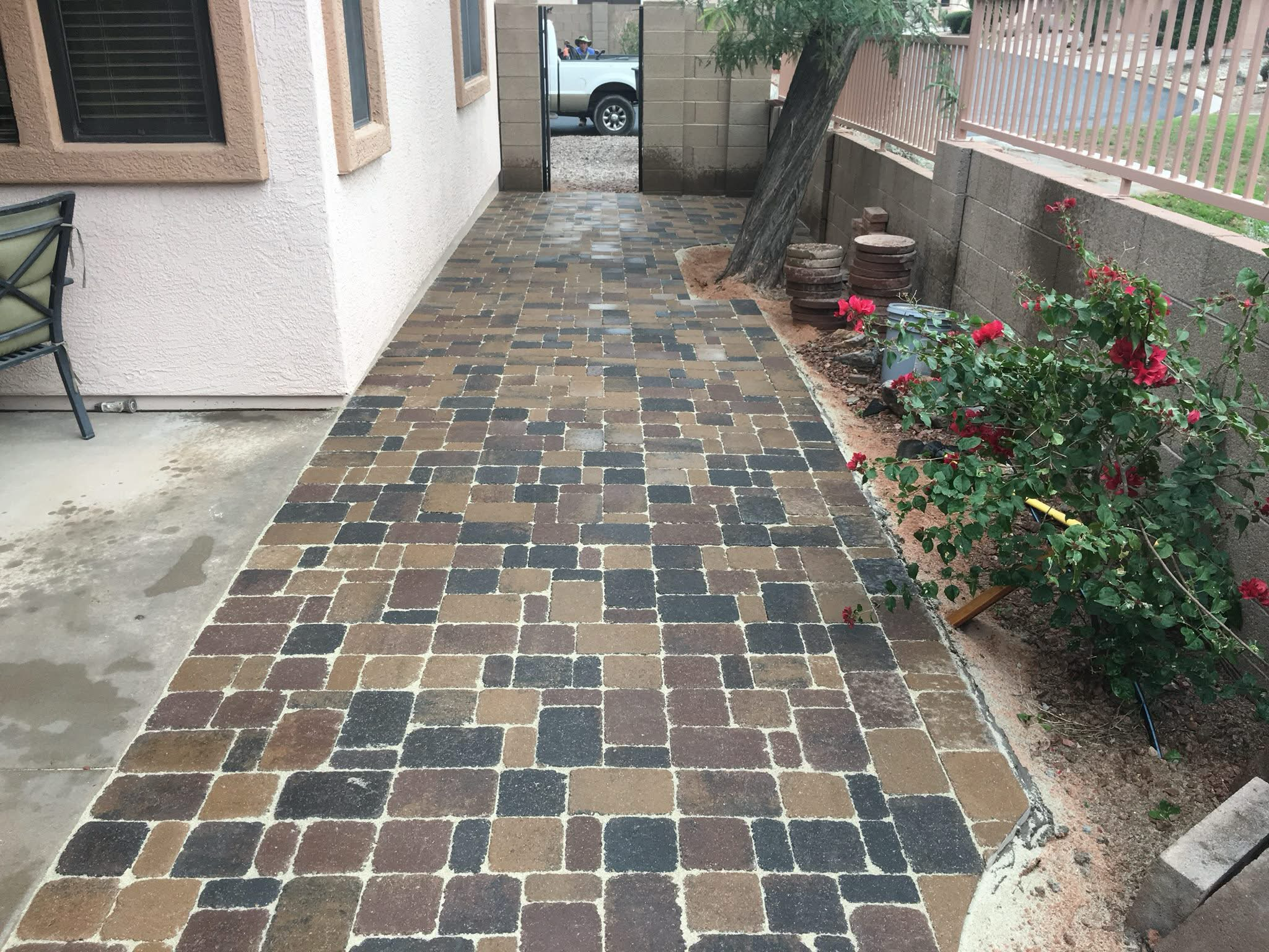 Outdoor brick patio with multicolored paver stones alongside a white stucco wall, with red flowering plants and trees near a pink fence.