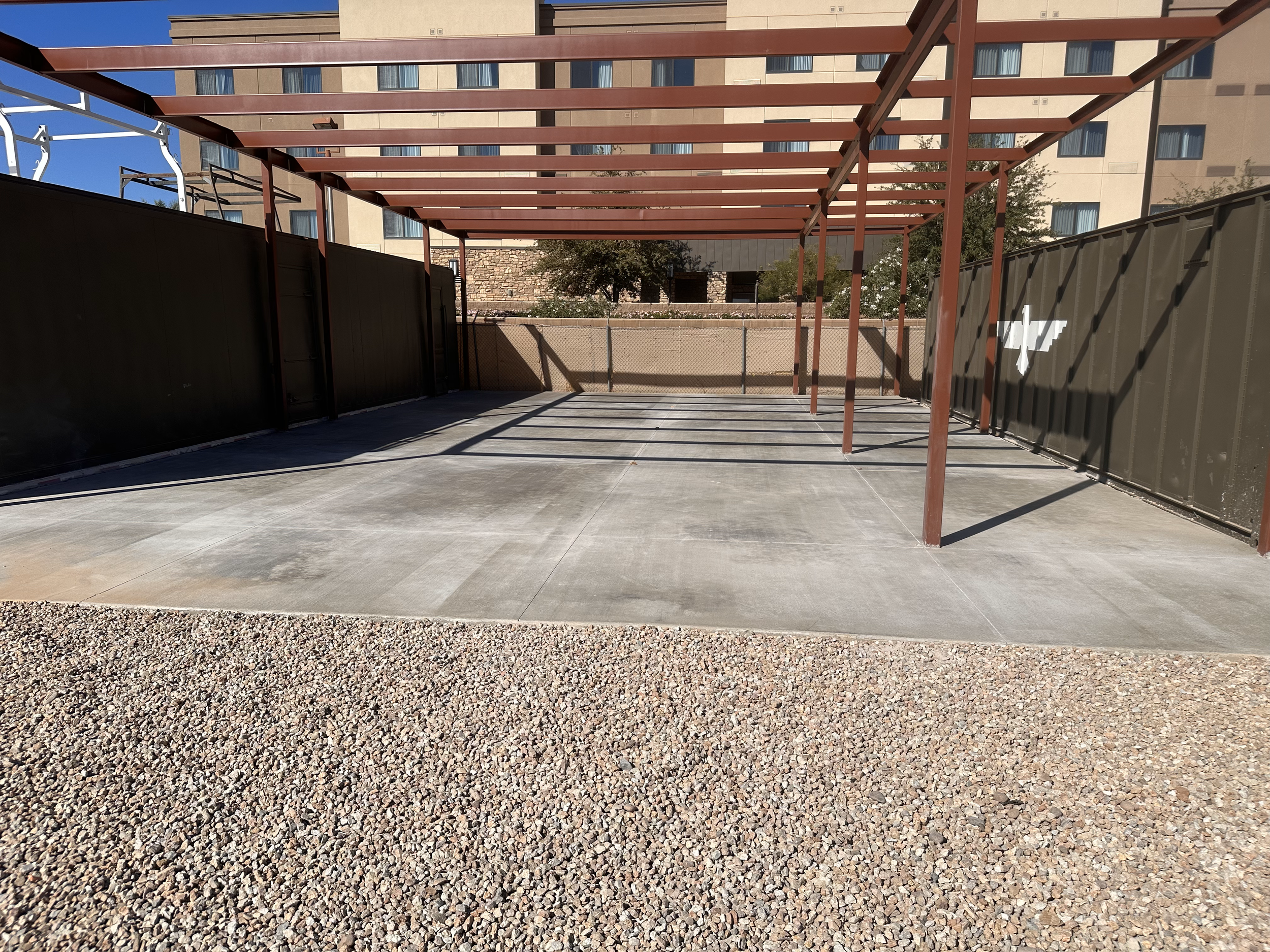 Empty outdoor space with a metal frame structure near buildings, featuring concrete flooring and gravel area, under blue sky.