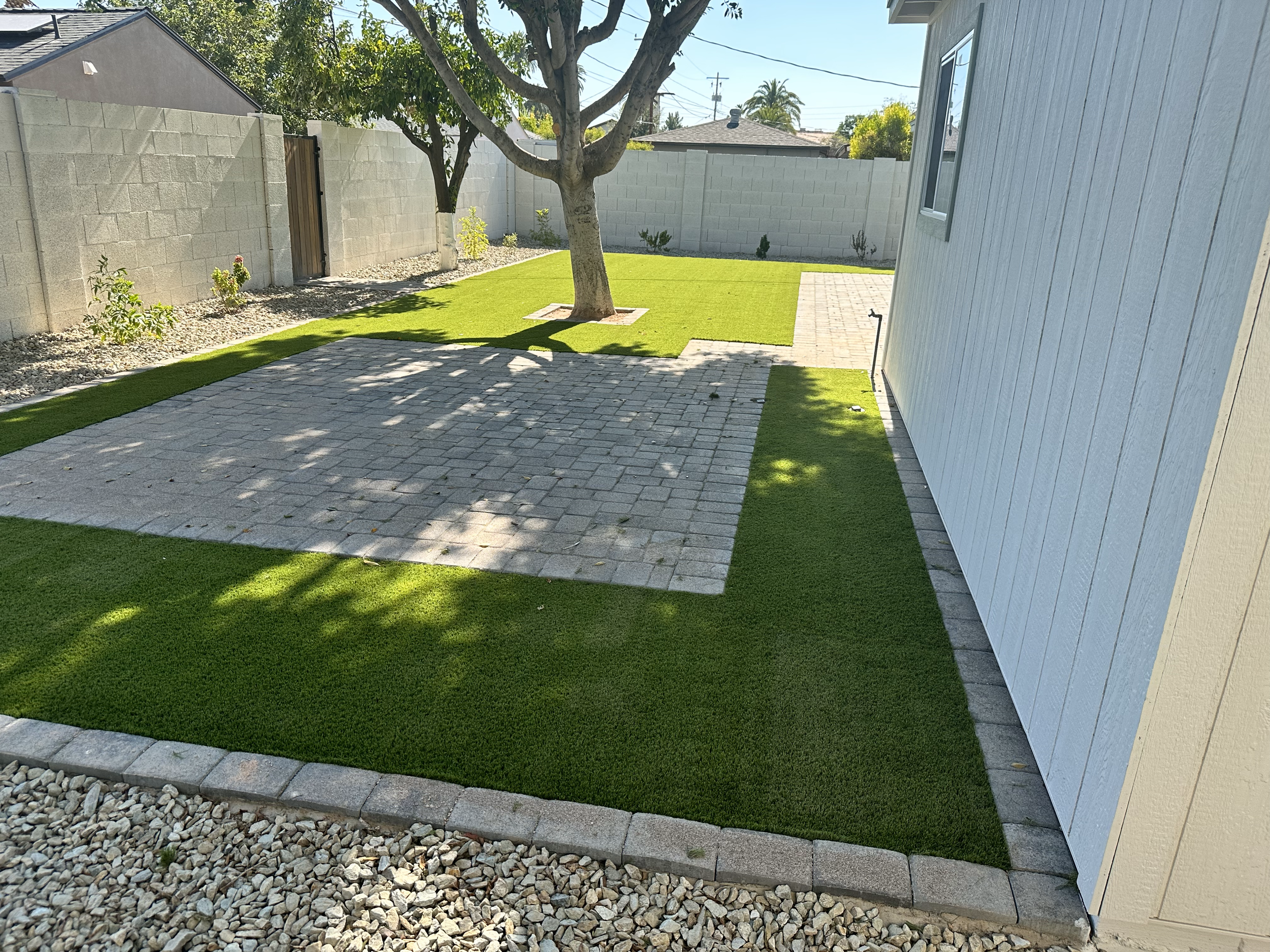 Backyard with artificial grass, a stone patio, and a tree in the center. A light-colored wall surrounds the yard. A small building is on the right with a window and light siding.