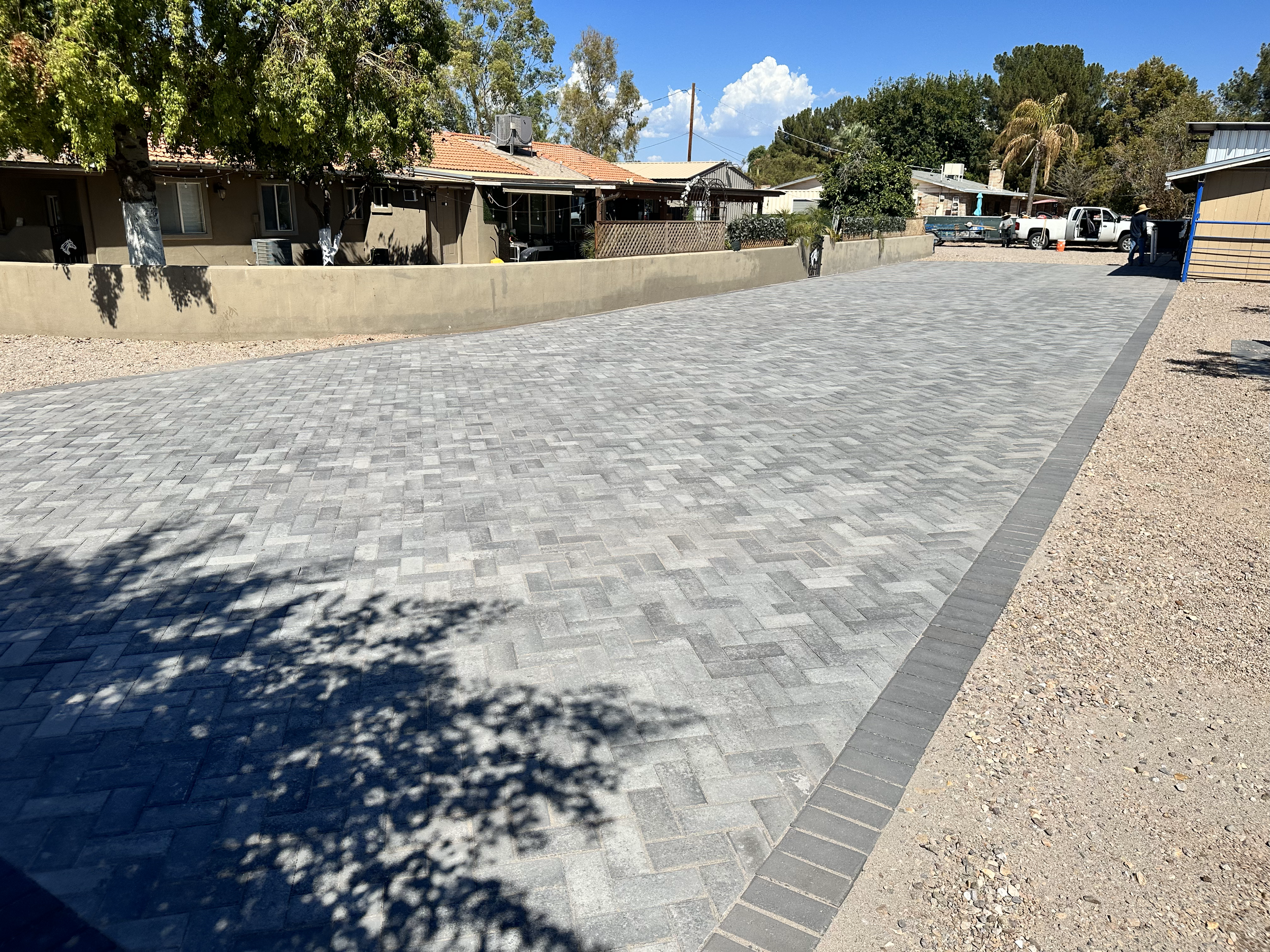 Large paved driveway with herringbone pattern surrounded by residential homes, trees, and gravel area under a clear blue sky.