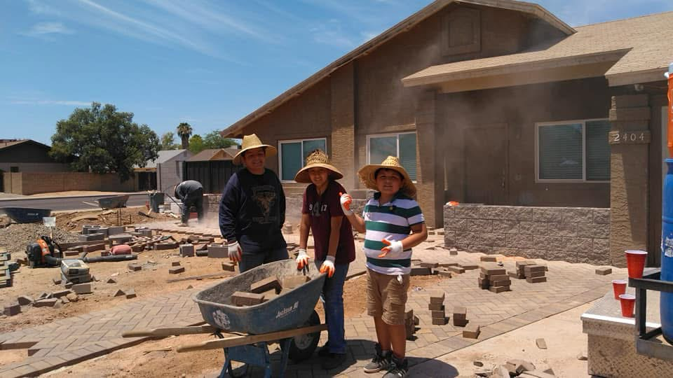 Three people wearing straw hats standing in front of a house under construction, holding construction materials near a wheelbarrow. The yard is filled with bricks and construction tools under a clear blue sky.
