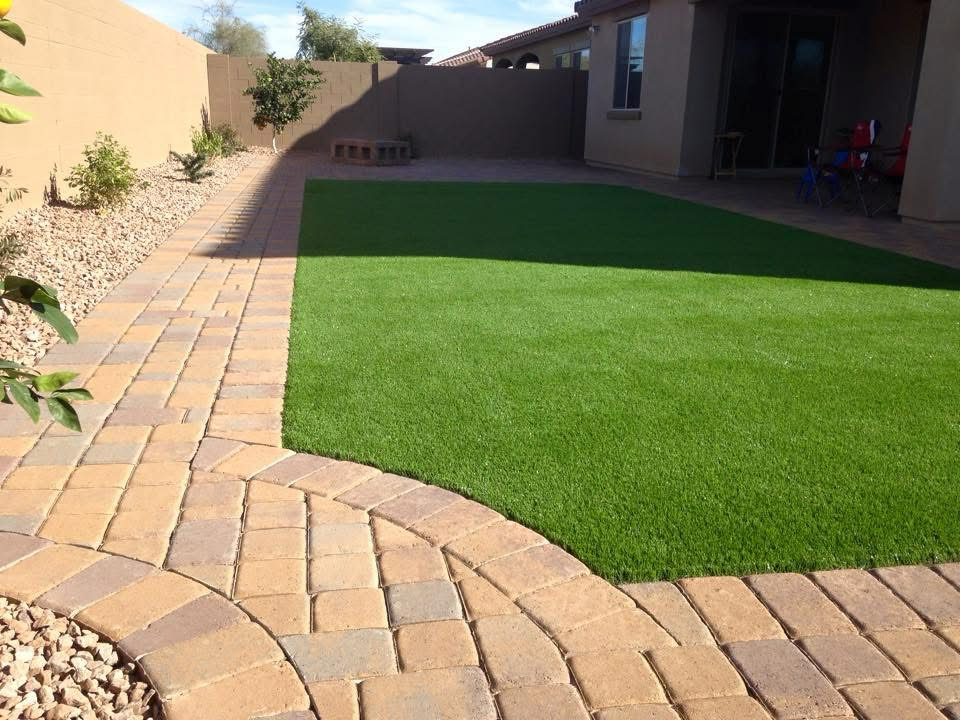 Backyard with artificial grass, brick patio, and desert landscaping including gravel and small trees along the edges.