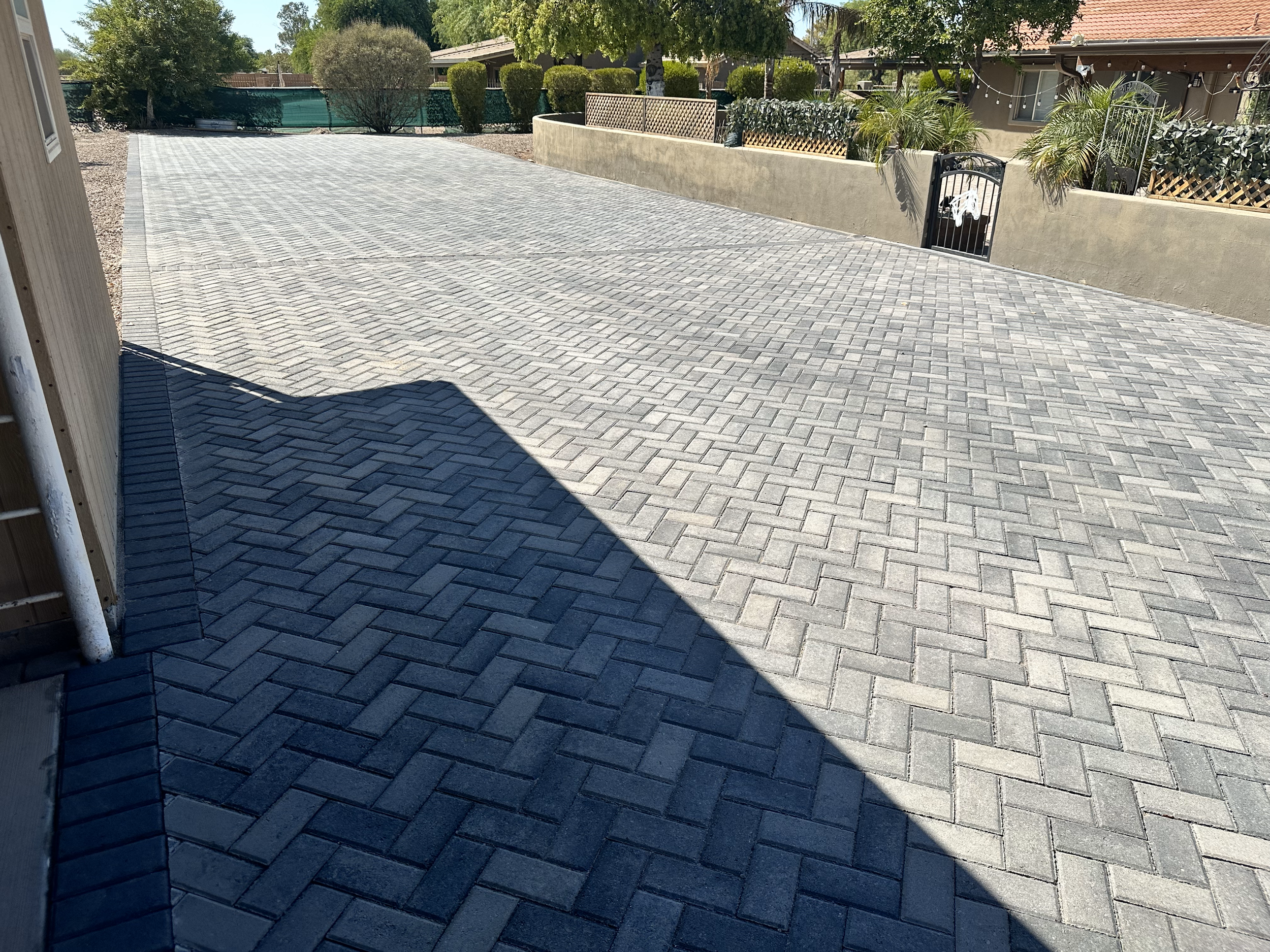 Brick driveway with herringbone pattern and shadows, surrounded by greenery and a gated entrance.