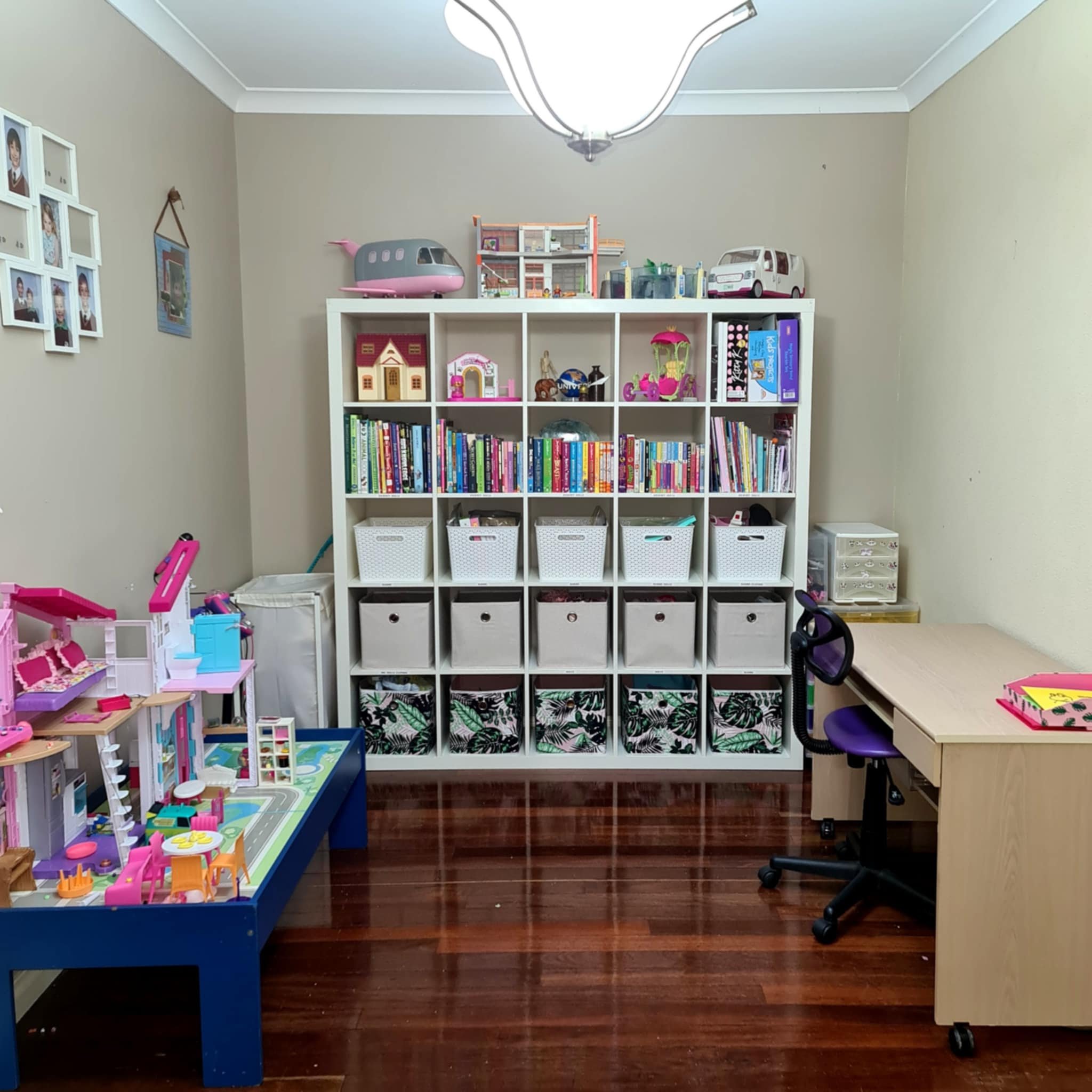 A room with a children's play area and storage. A white shelving unit holds books, toys, and storage baskets. A play table with a toy set is on the left. A desk with a chair is on the right, under bright lighting. Walls have framed photos.