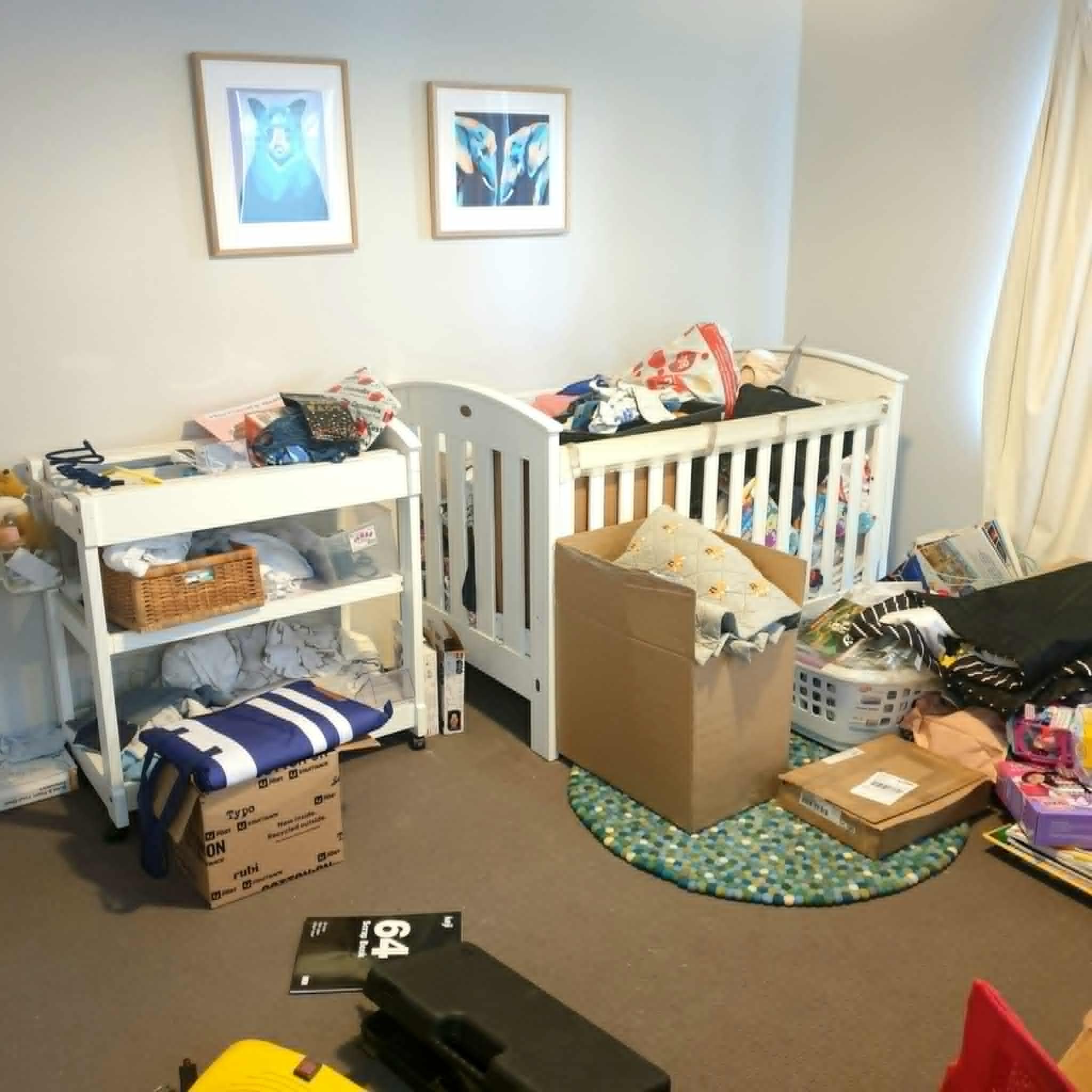Messy nursery with crib, changing table, boxes, laundry basket, and assorted items; framed animal prints on wall.