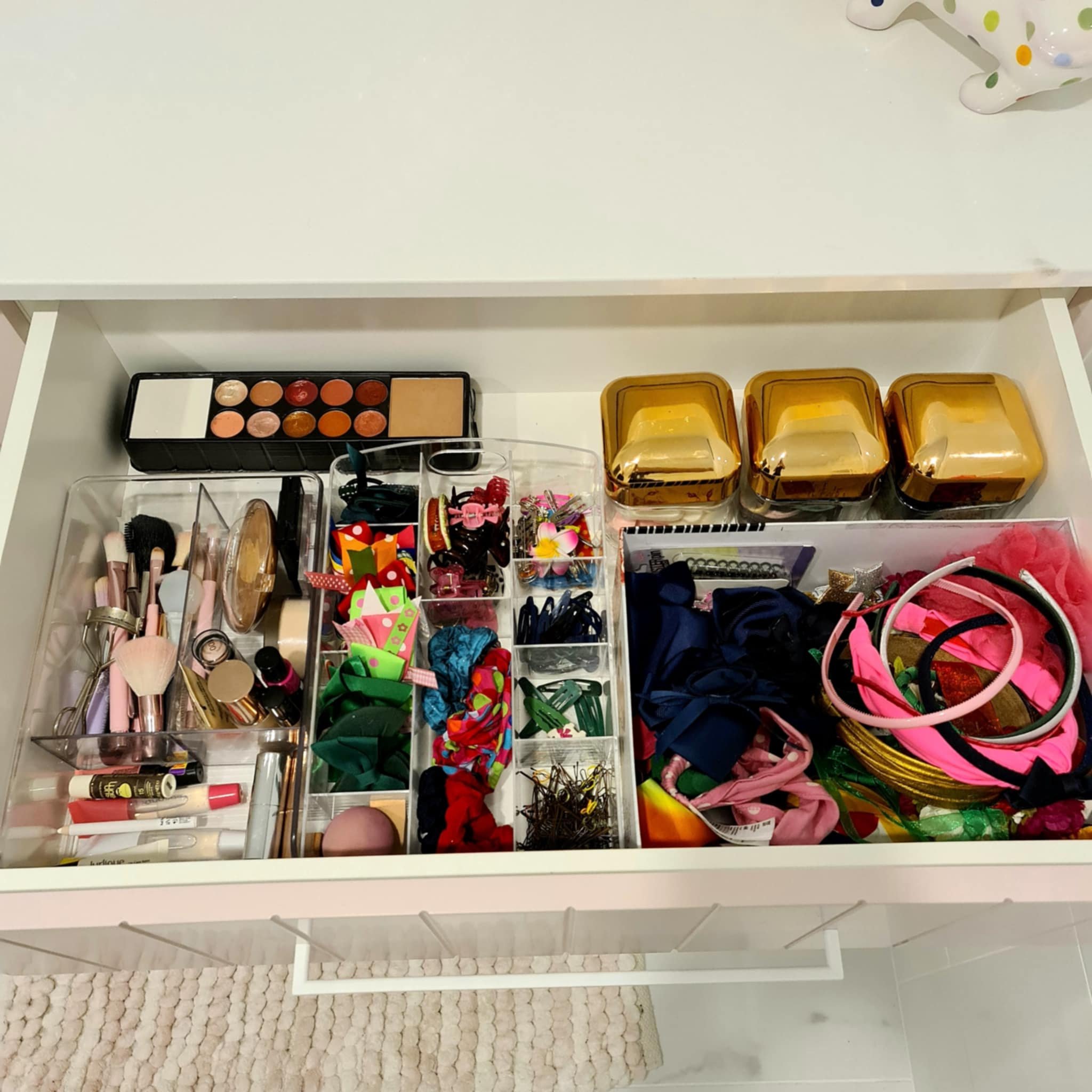 Drawer organized with makeup products, hair accessories, and brushes; includes eyeshadow palette, makeup brushes, headbands, and small containers for clips and scrunchies.