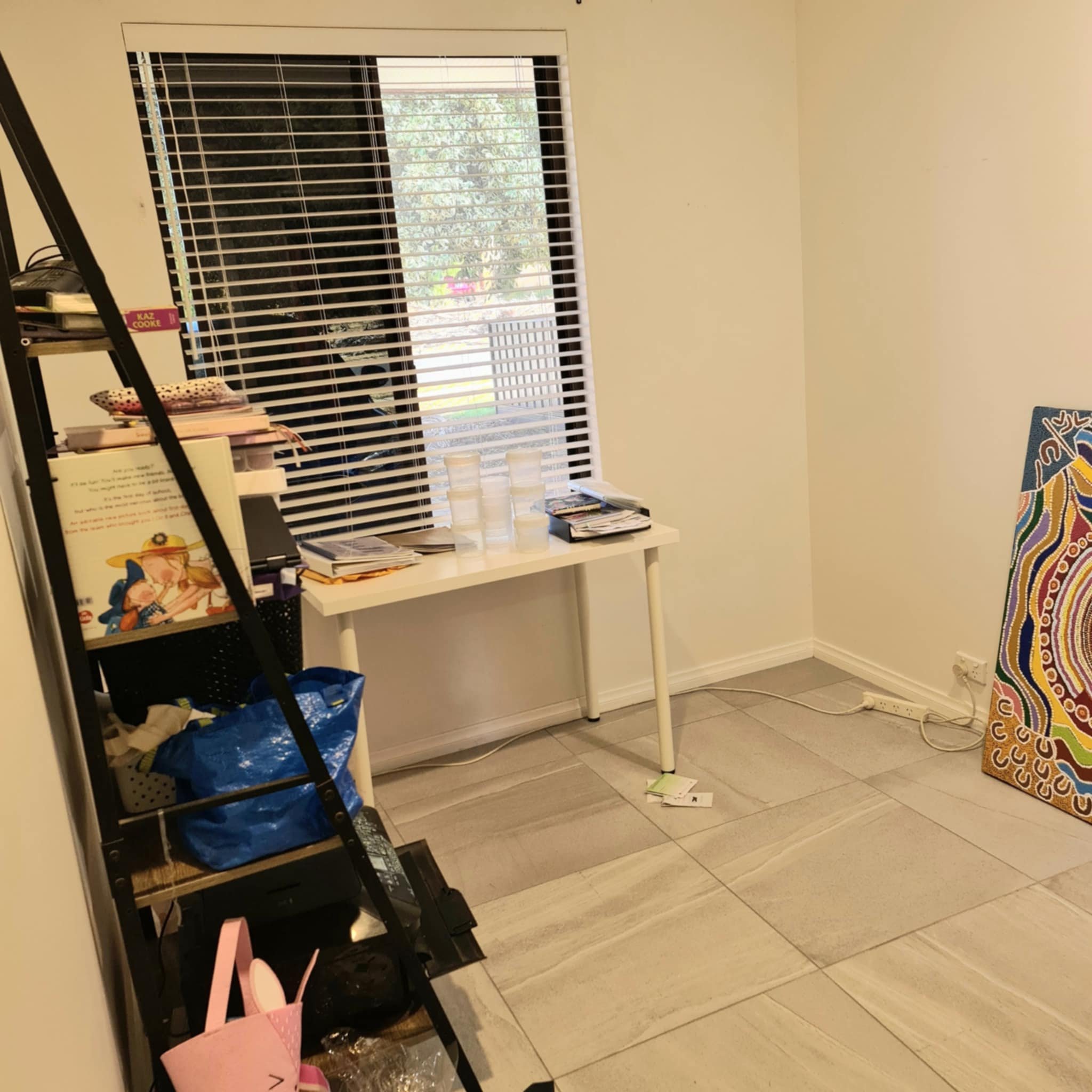 Small room with a white table under a window with blinds, surrounded by scattered books, papers, and art supplies. A shelving unit holds various items. A large colorful painting leans against the wall near the corner.