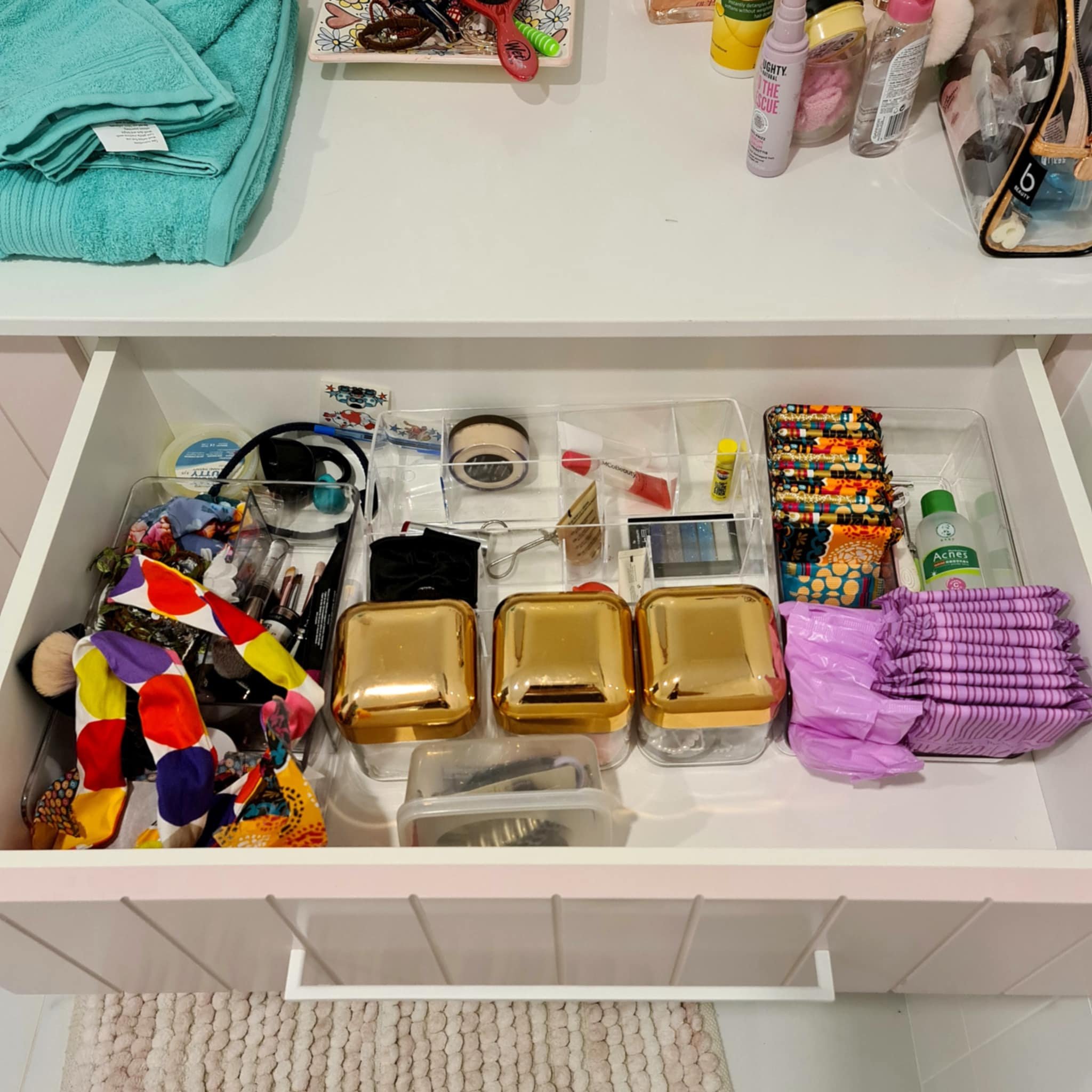 Open drawer containing well-organized personal care and beauty items in clear organizers, including makeup, hair accessories, skincare products, and feminine hygiene products. Above the drawer, towels and toiletries are placed on a countertop.