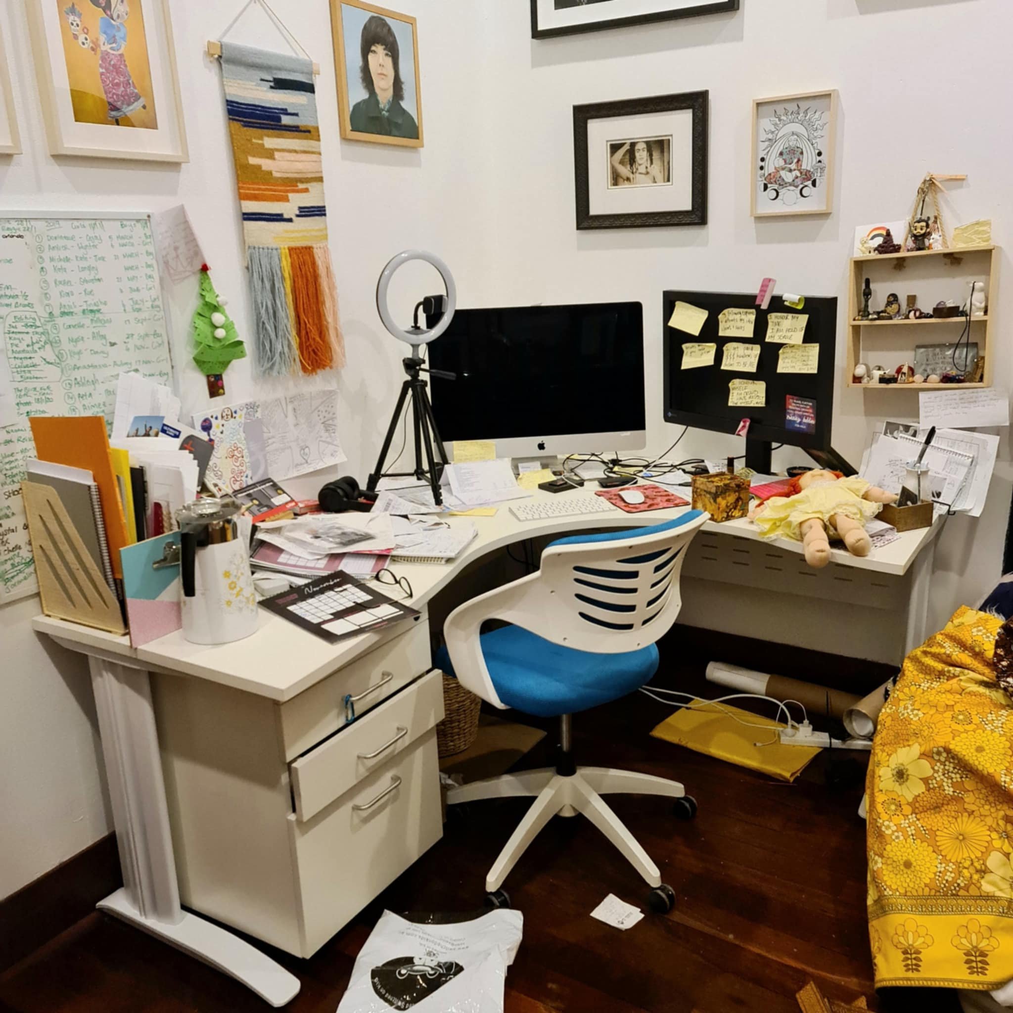 A cluttered home office space with a white desk and chair, computer monitors surrounded by sticky notes, and various office supplies and papers scattered. Walls have colorful artwork and photographs. A ring light is set up next to the monitor, and sh