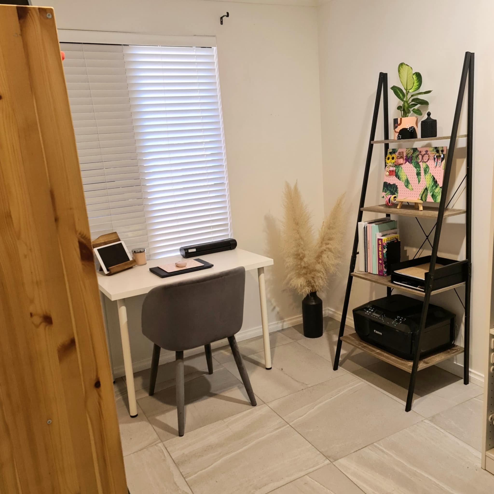 Small minimalist home office setup with a white desk, gray chair, tablet, and laptop. A black ladder shelf holds books, a printer, and decorations. Blinds cover the window and a plant decorates the room.