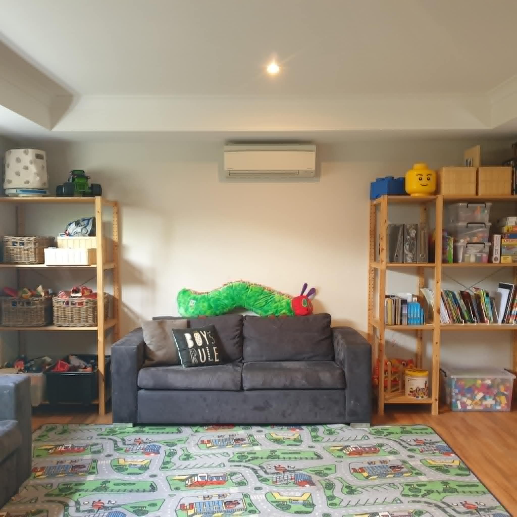 Children's playroom with shelves, toys, books, and a gray sofa. A colorful caterpillar plush is on the sofa, and a road-themed play rug covers the floor.