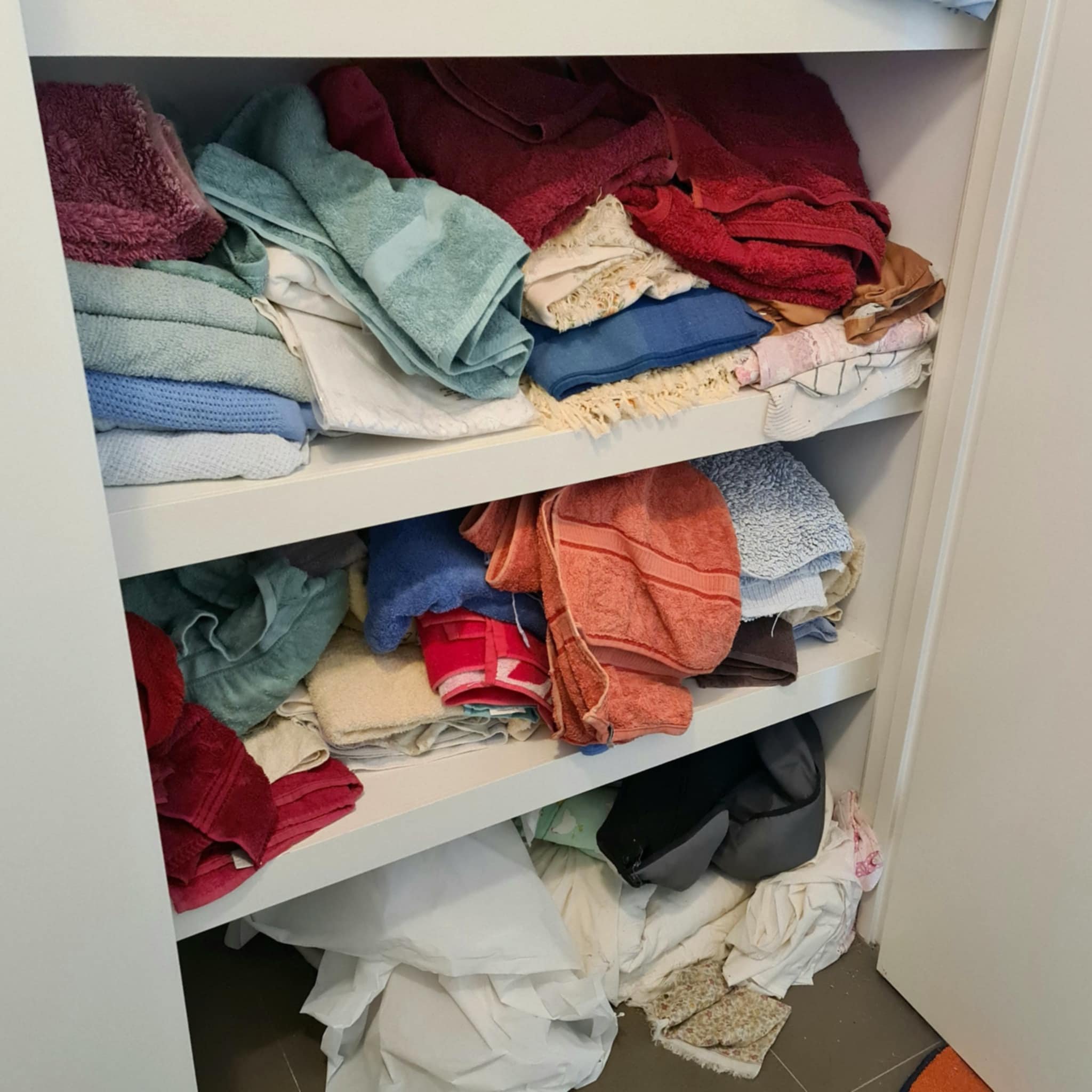 A closet with neatly stacked towels and linens on shelves. Various colors of towels are visible, including red, blue, green, and orange. On the bottom shelf, there are some crumpled white fabrics and a small black bag.