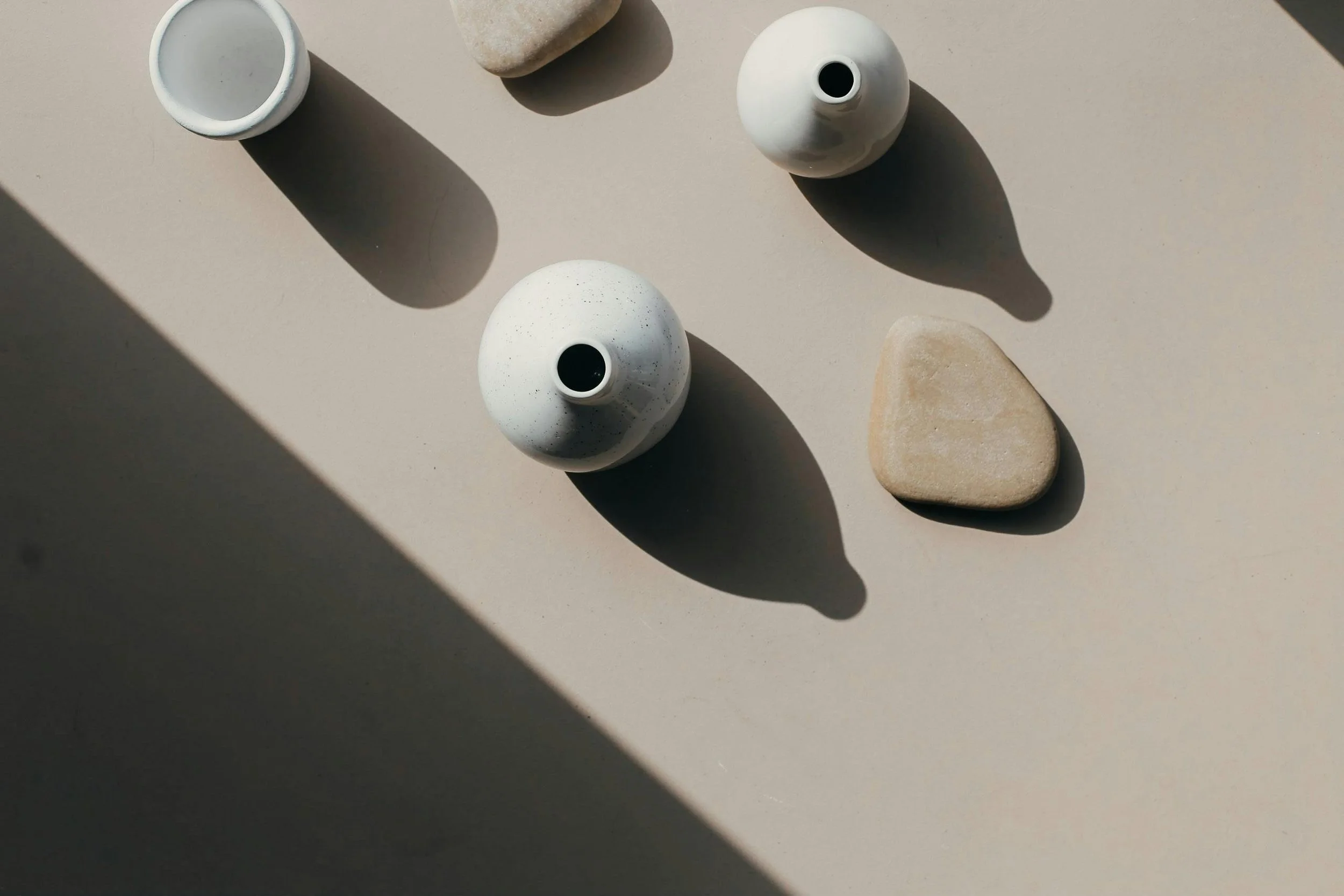 A top-down view of four objects on a light-colored surface, including a small white bowl, rounded stones, a white vase, and a flat piece of stone, casting shadows in the sunlight.