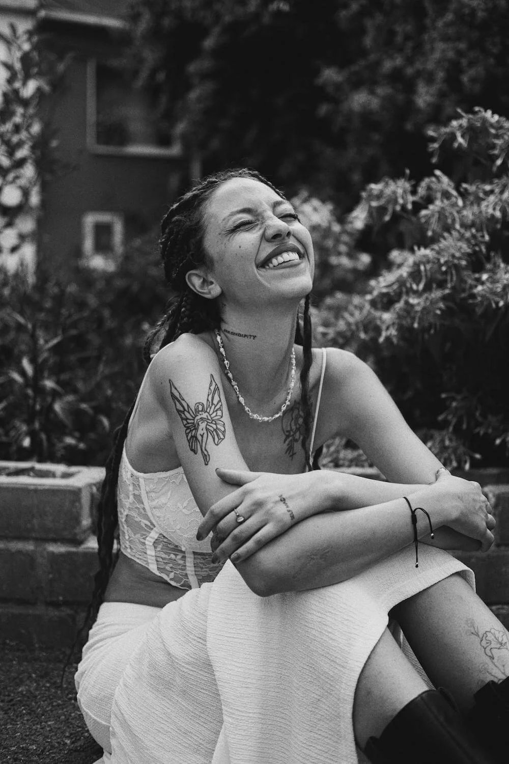 A woman with long dark hair styled in braids, wearing a white lace top and a white skirt, sitting outdoors surrounded by plants. She has tattoos on her arm and neck, and is laughing with her eyes closed.