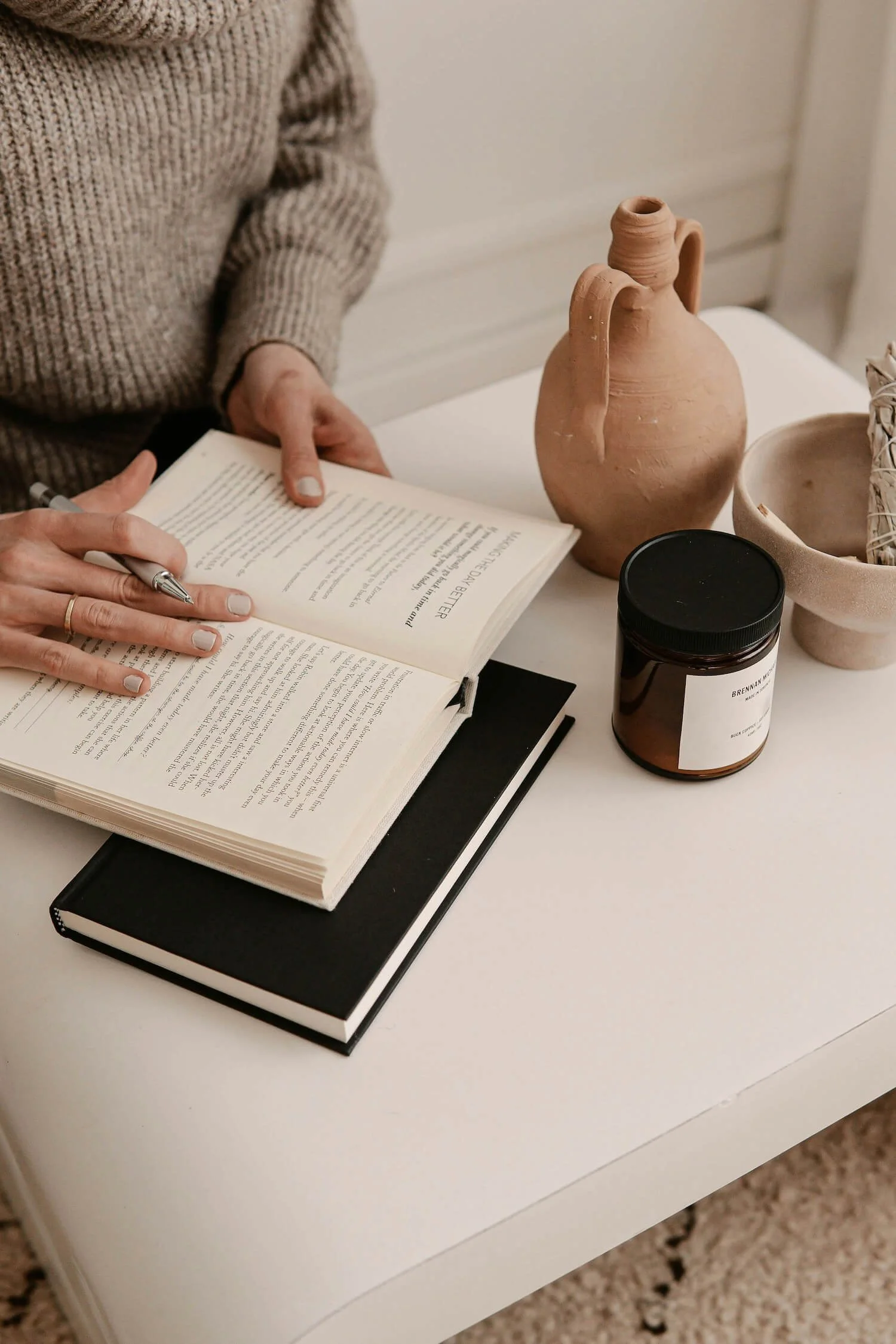 A person in a beige sweater is writing in an open book with a pen. The book is on top of a black notebook, on a white table. There are decorative items on the table including a terracotta vase, a jar, and a bowl with woven items inside.