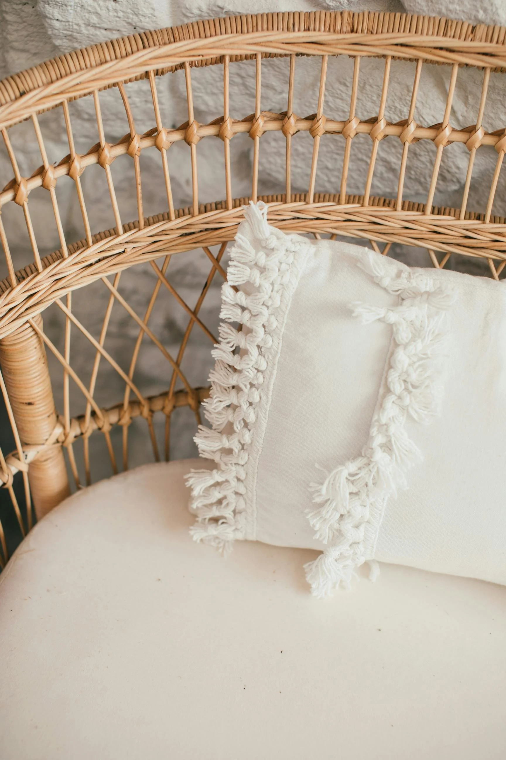 A cream-colored pillow with fringe trim on a light-colored sofa next to a wicker chair with a textured stone wall in the background.