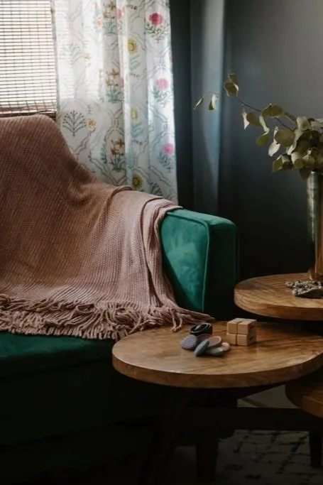 Cozy living room corner with a green velvet armchair, pink throw blanket, wooden side table with speckled stones and wooden cube, and a tall vase with leafy branches near window with floral and lace curtains.