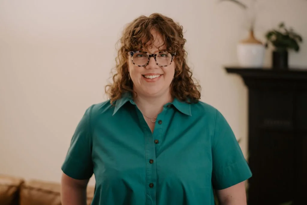 A woman with curly red hair, glasses, and a teal shirt smiling indoors with a white wall, a dark fireplace, and potted plants in the background.