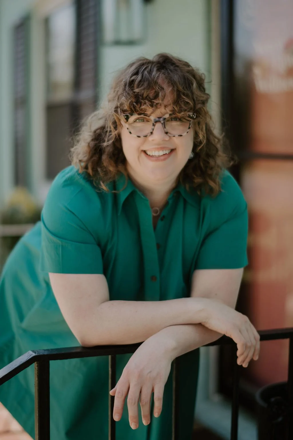 A woman with curly brown hair and glasses smiling, wearing a green shirt, outdoors with a blurred background.