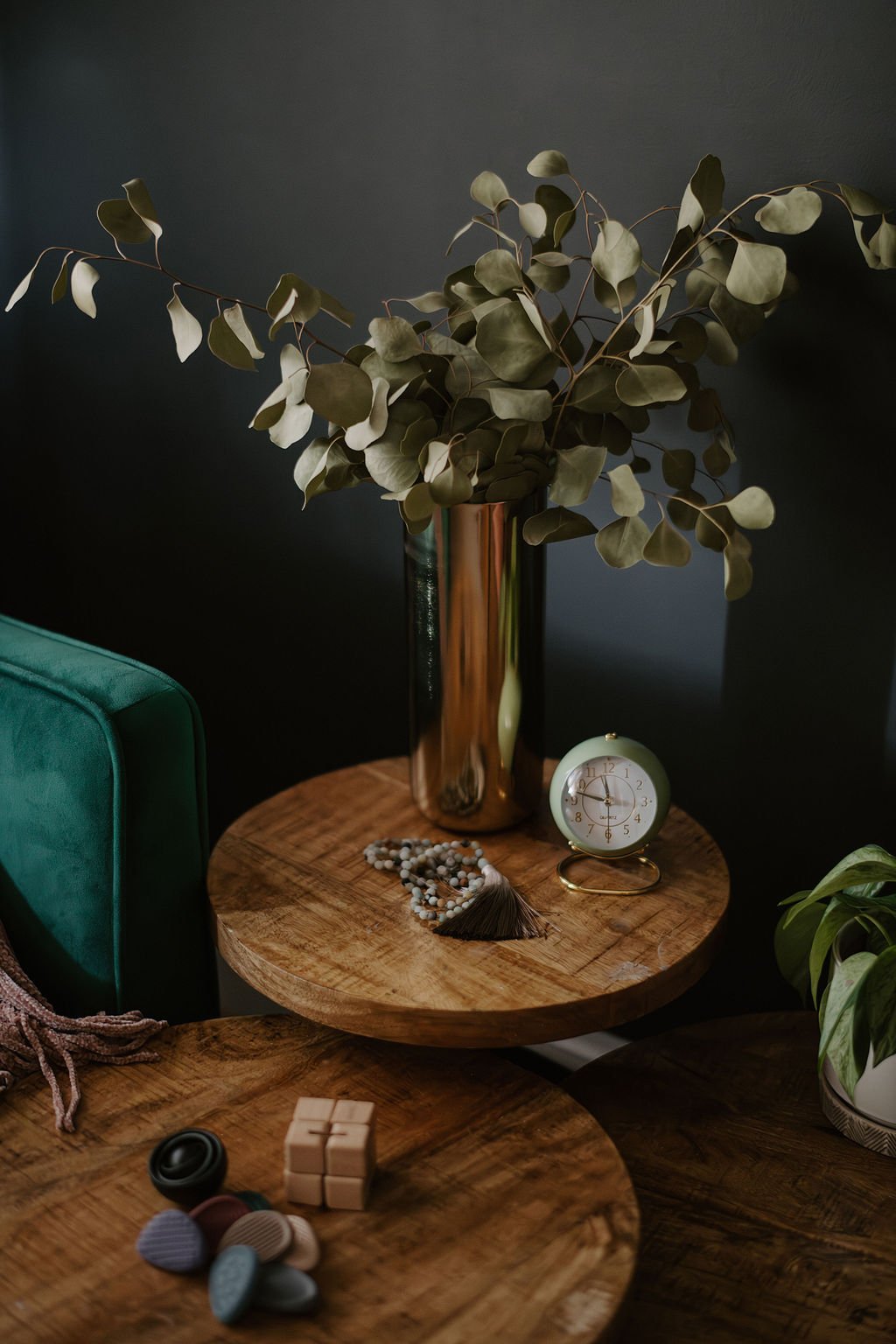 A wooden table with a tall metallic vase holding dried eucalyptus branches, a small clock, a beaded necklace with a tassel, and a green potted plant in a white pot, placed against a dark background.
