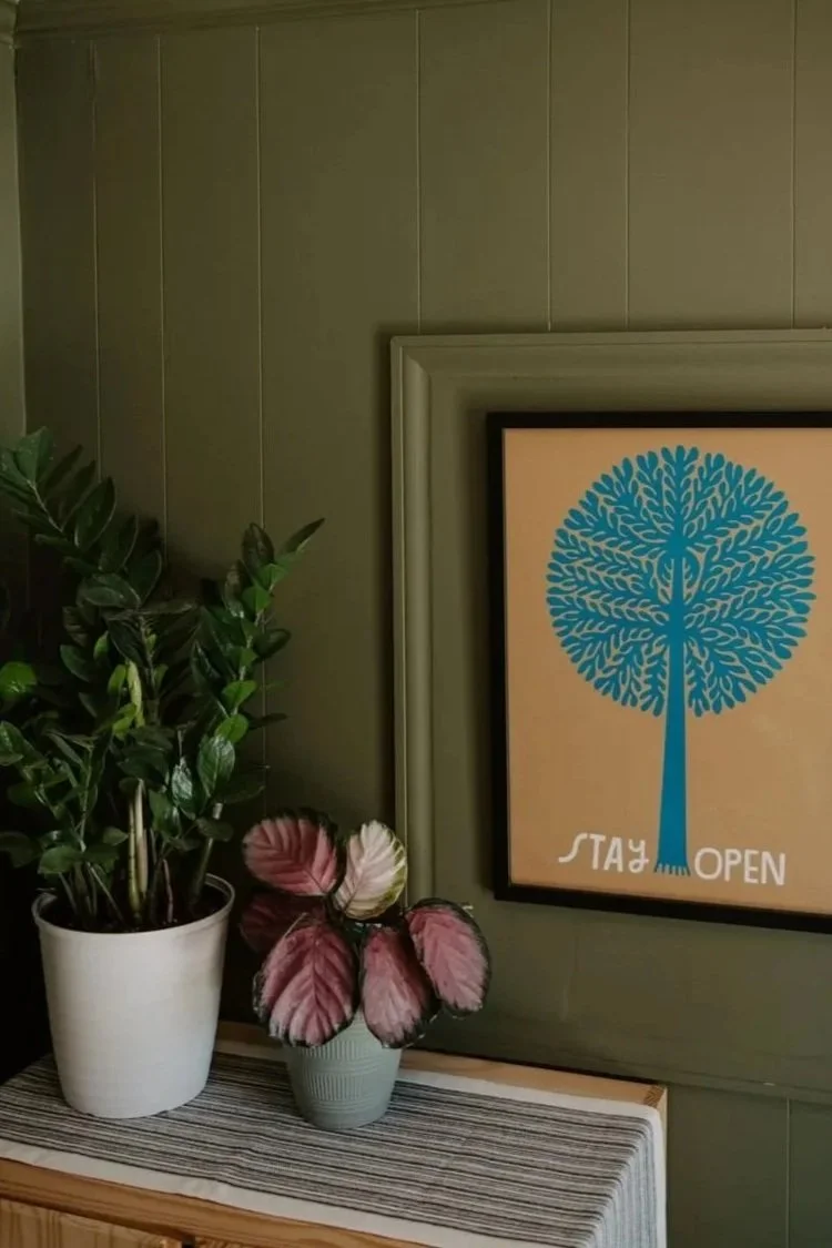 Interior with two potted plants next to a framed sign with a blue tree and the text "STAY OPEN" on a sage green wall.