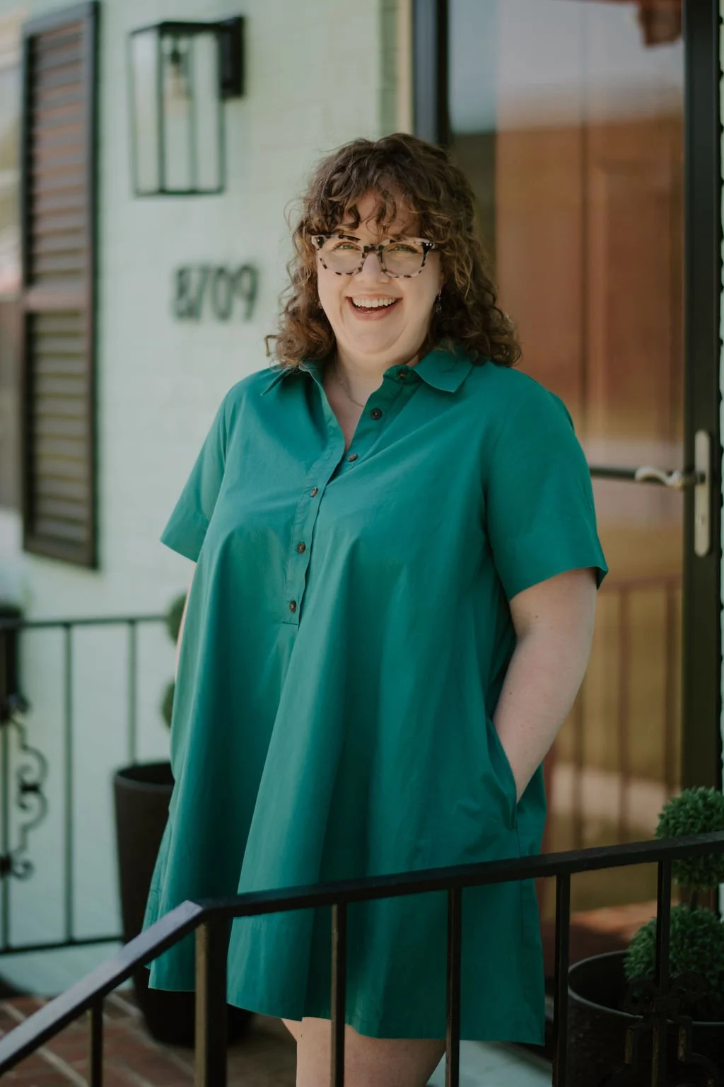 A woman with curly brown hair, wearing glasses and a teal dress, smiling and standing outside a house.