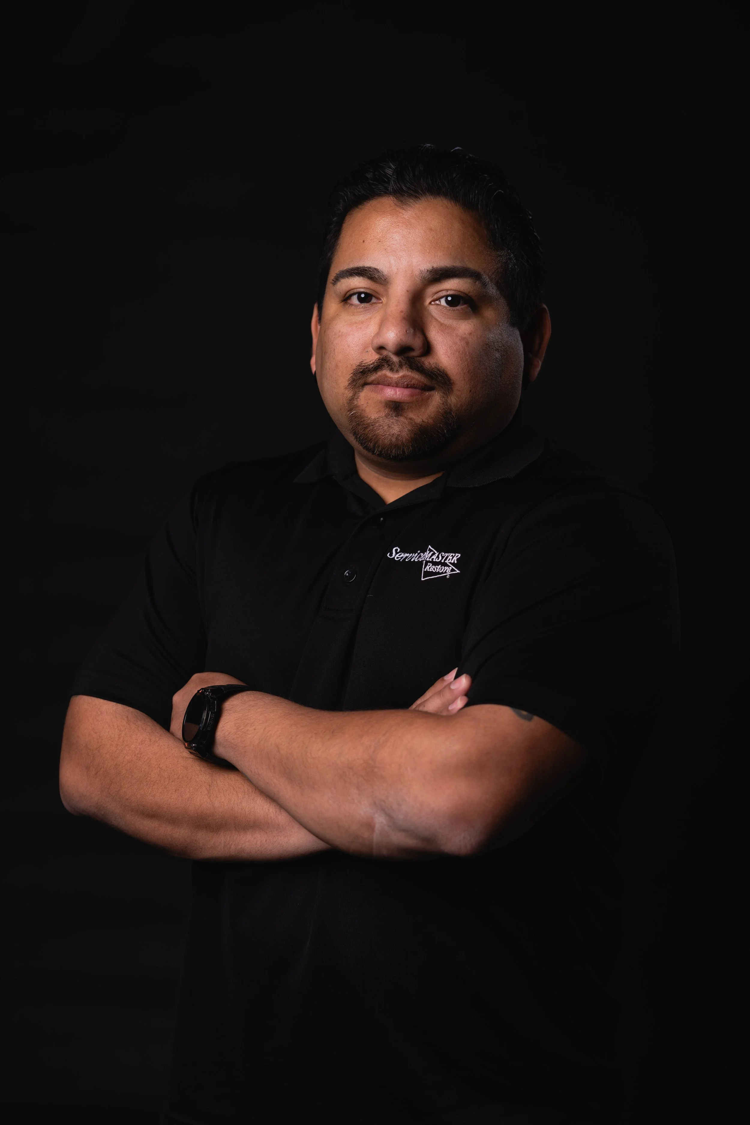 A man smiling with arms crossed, wearing a black collared shirt with a logo that says 'Service Master Restore' on a black background.