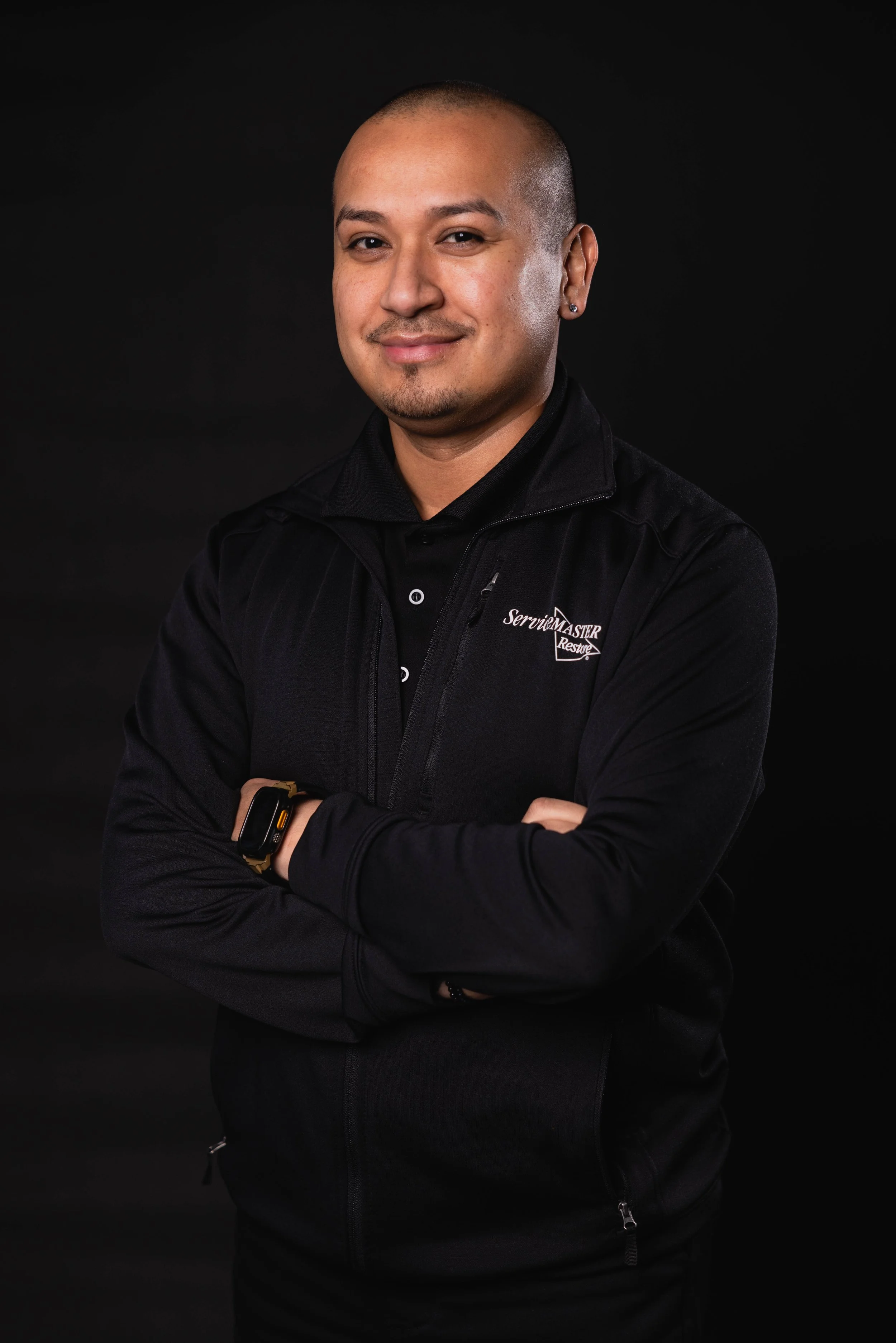 A man smiling with arms crossed, wearing a black jacket with a logo that says 'Service Master Restore' on a black background.