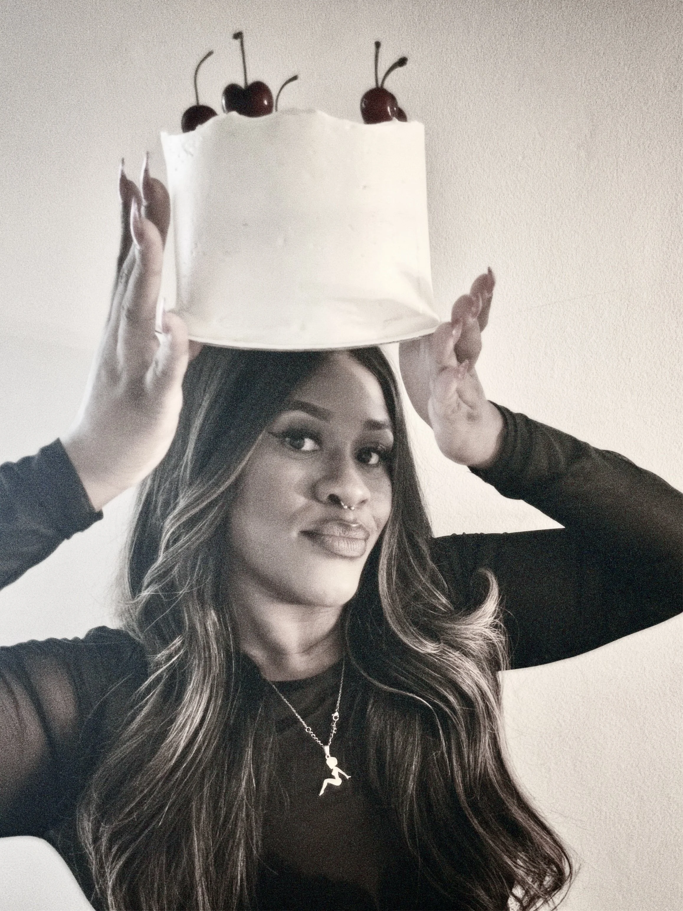 A woman with long hair and a septum piercing is holding a white cake decorated with cherries on top, above her head.