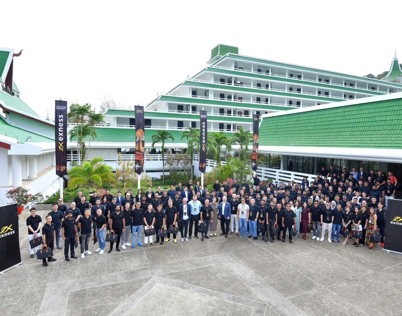 Group photo of many people standing outdoors in front of a large modern building with green roofs, palm trees, and banners for 'exness' during daylight.