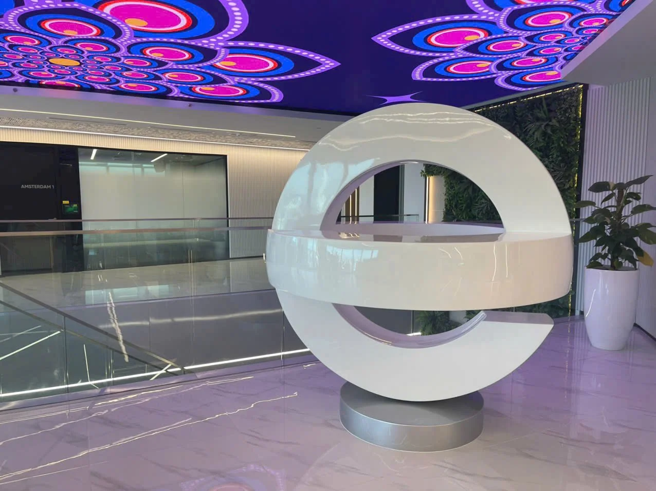 Modern interior space with colorful, purple-lit patterned ceiling, a futuristic white sculpture resembling a ring or portal, glass railing, and a potted plant.