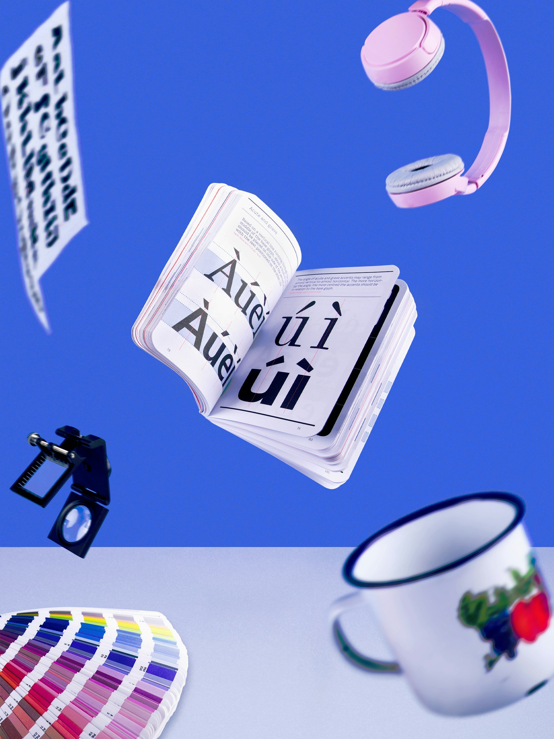 Floating items including an open book with typography, pink headphones, a paint color palette, a paint mug, a magnifying glass, and a speech bubble against a blue background.