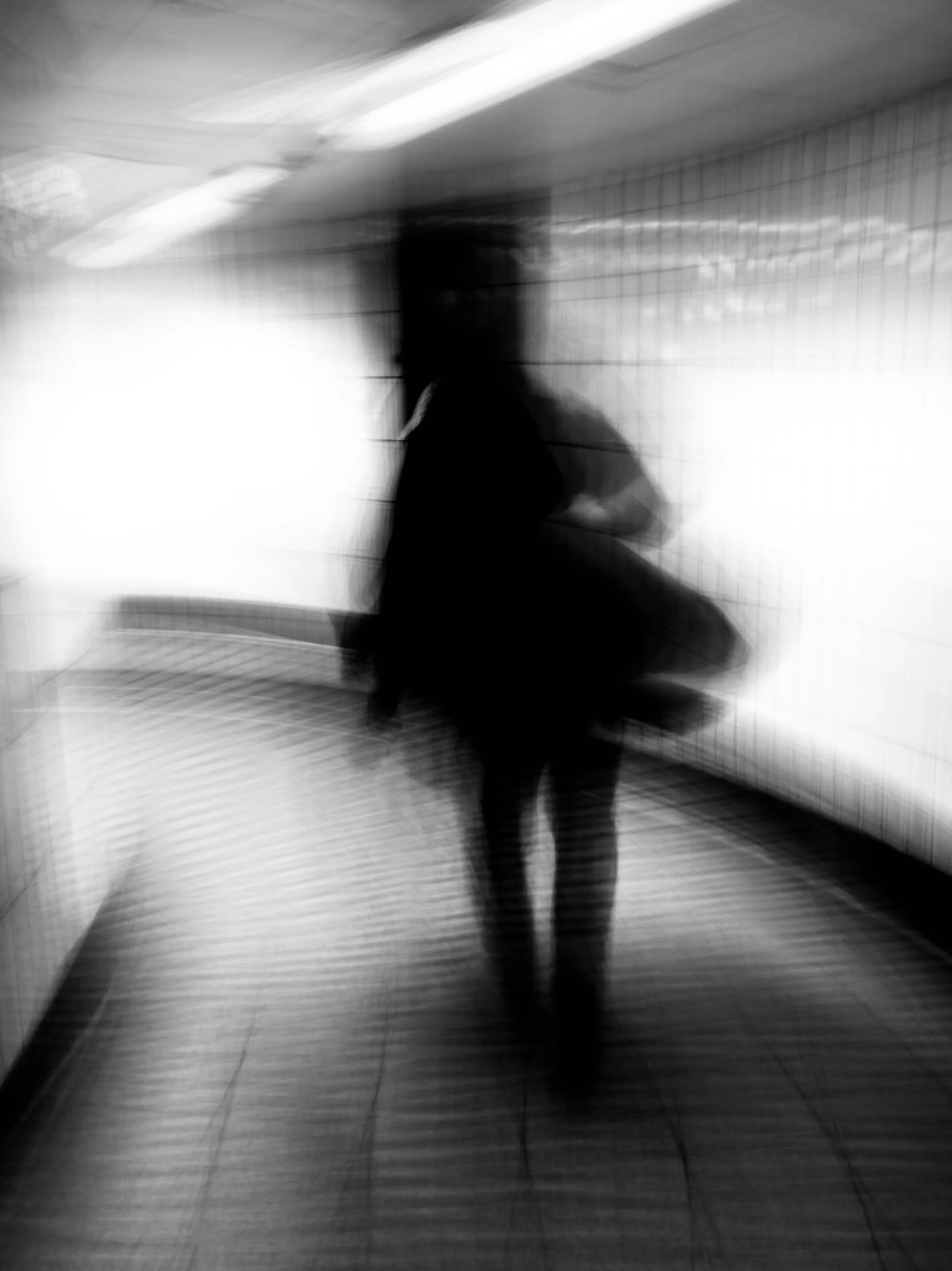 Black and white photo of a person walking in a corridor with tiled walls, blurred and out of focus.