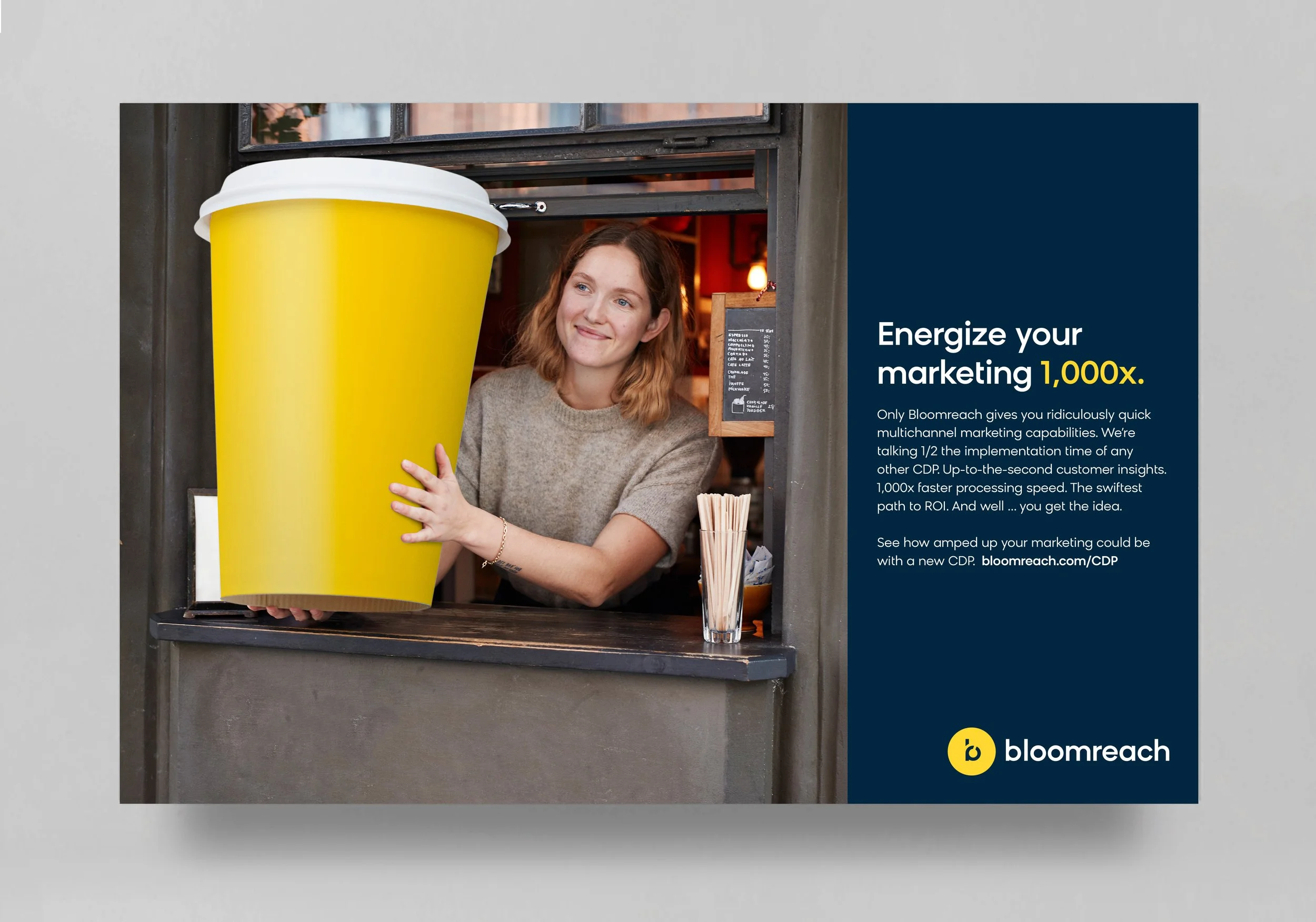 A woman in a beige sweater is smiling and holding a large yellow coffee cup at a small cafe window. There is a small chalkboard menu and a glass of stir sticks on the counter.