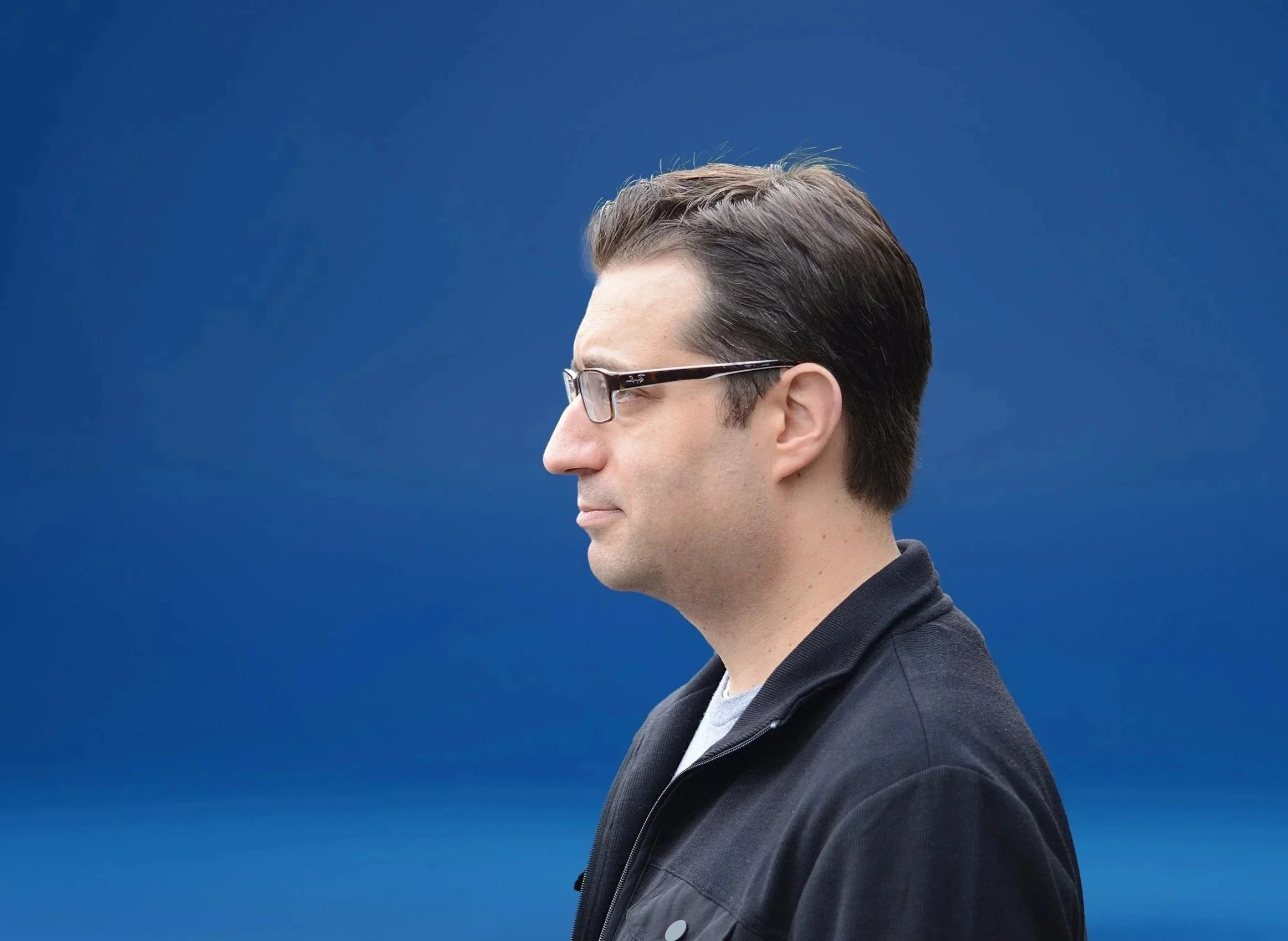 Profile side view of a young man with brown hair, wearing glasses, a dark jacket, and a light-colored shirt, against a blue background.