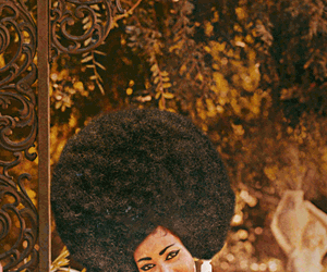 A woman with a large afro hairstyle smiling outdoors with trees and decorative elements in the background.