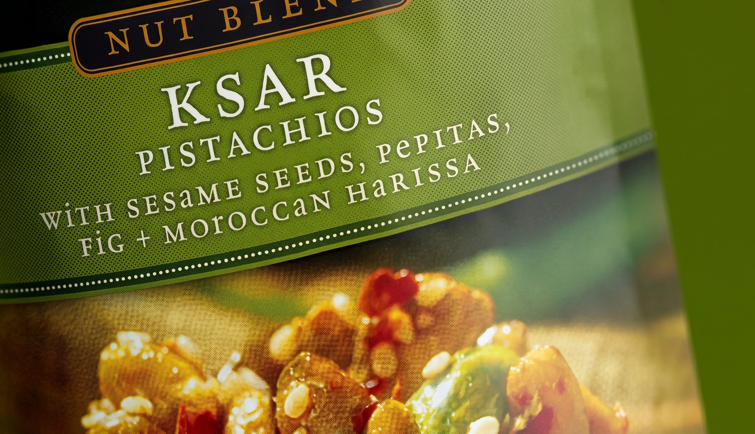 Close-up of a food packaging label for nut blend pistachios, with sesame seeds, pepitas, fig, and Moroccan harissa, featuring green background design and an image of the snack below.
