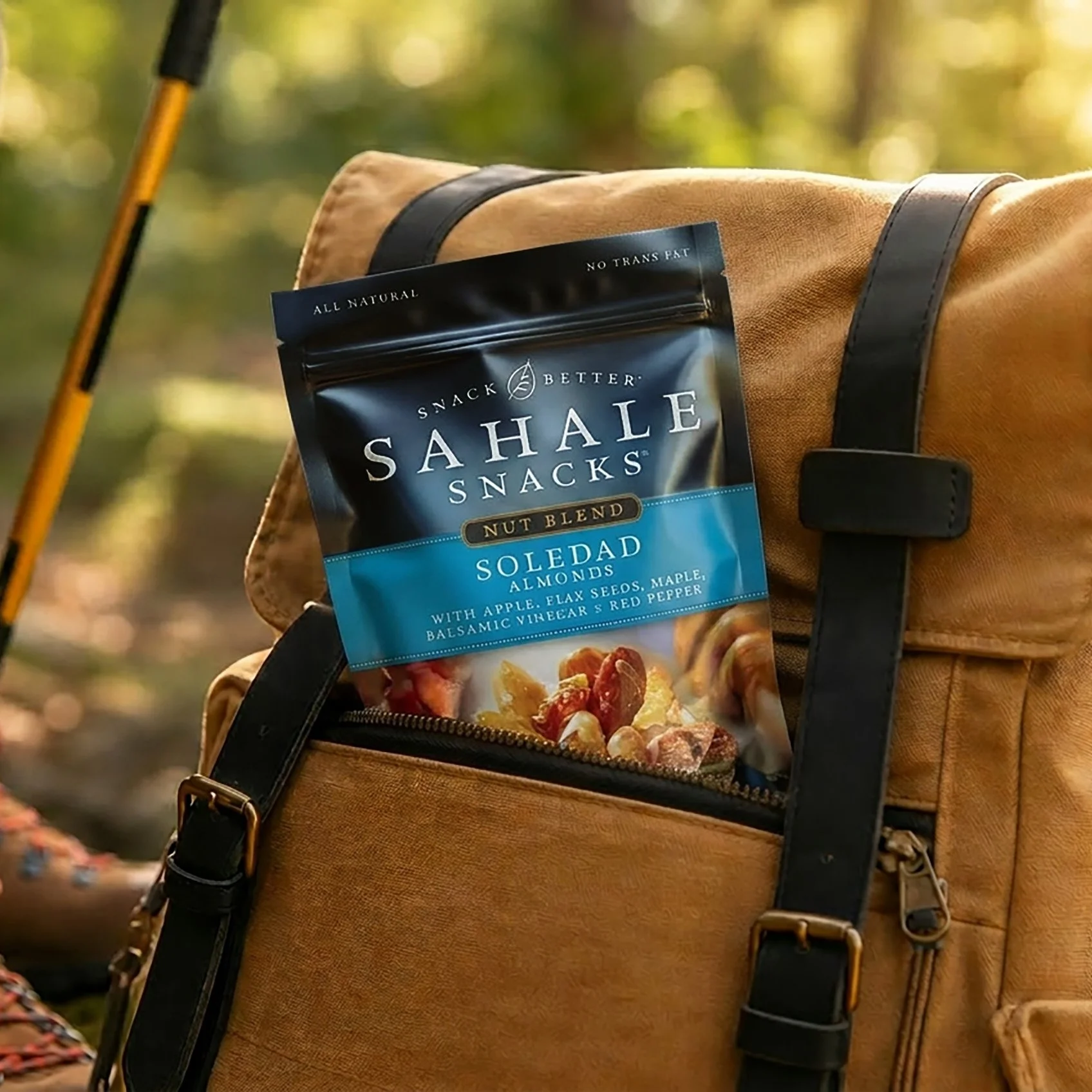 A backpack with a snack packet attached to the front pocket, set in an outdoor forest setting with yellow and green foliage.