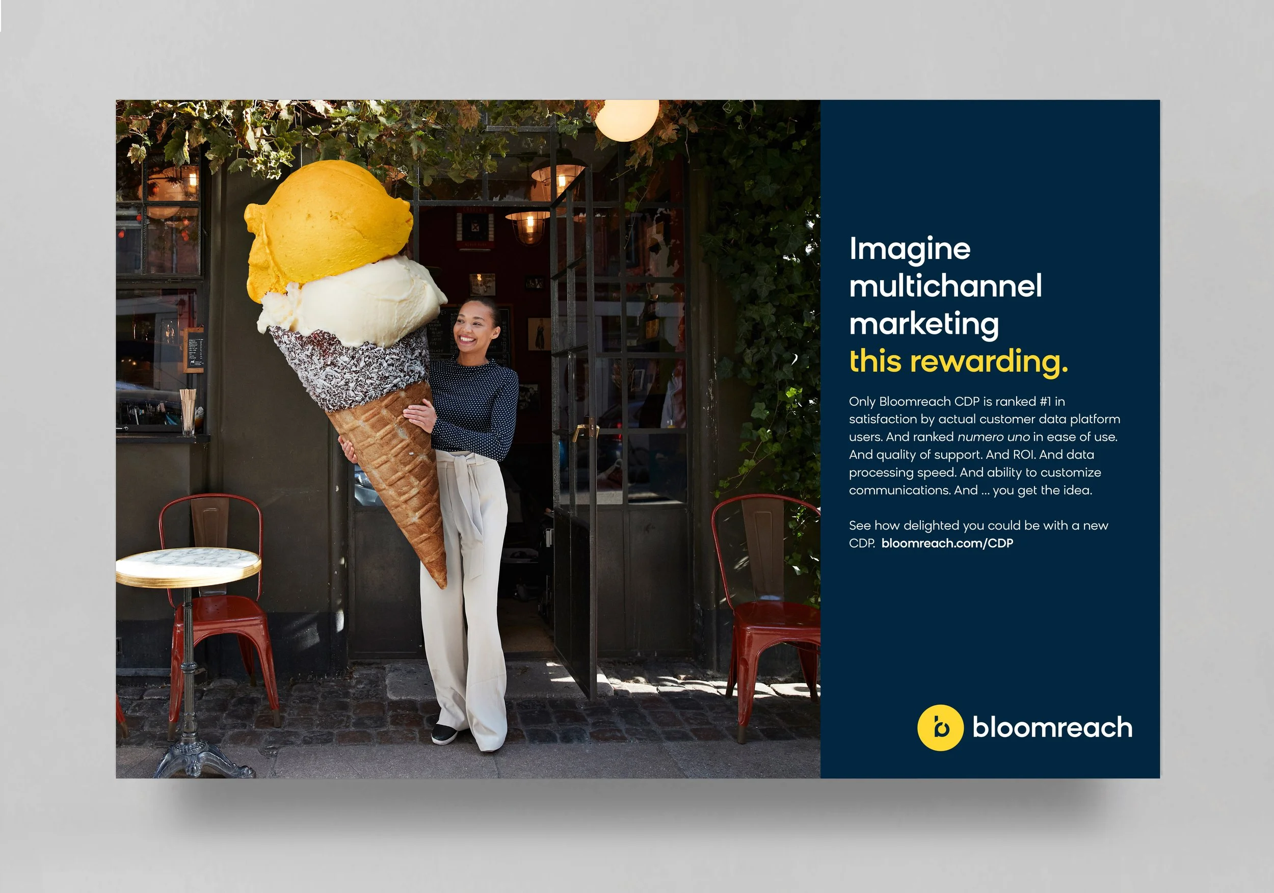 A woman standing in front of a cafe holding an oversized ice cream cone with yellow and white scoops. The cafe has a dark exterior with greenery and orange chairs outside, and the woman is smiling and dressed in a polka dot top and light-colored pant