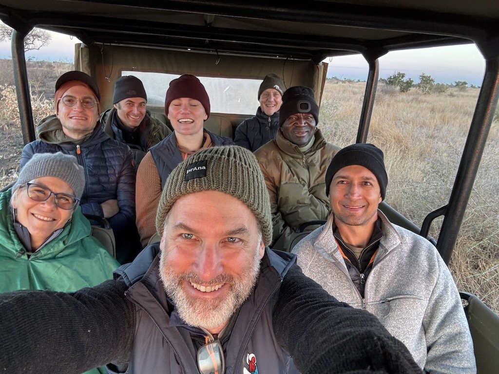 A group of people sitting in a room with wooden decor, engaged in a discussion. A man stands by a flip chart, presenting to the group. Chairs are arranged in a circle, and a beer bottle is on one of the side tables. Windows let in natural light.