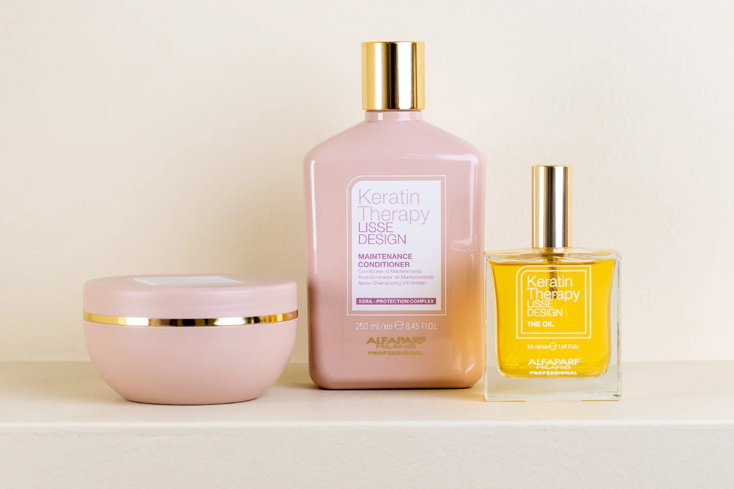 Pink container with gold trim, pink bottle labeled 'Keratin Therapy Lisse Design Maintenance Conditioner,' and yellow oil bottle labeled 'Keratin Therapy Lisse Design The Oil' displayed on a white shelf against a plain wall.