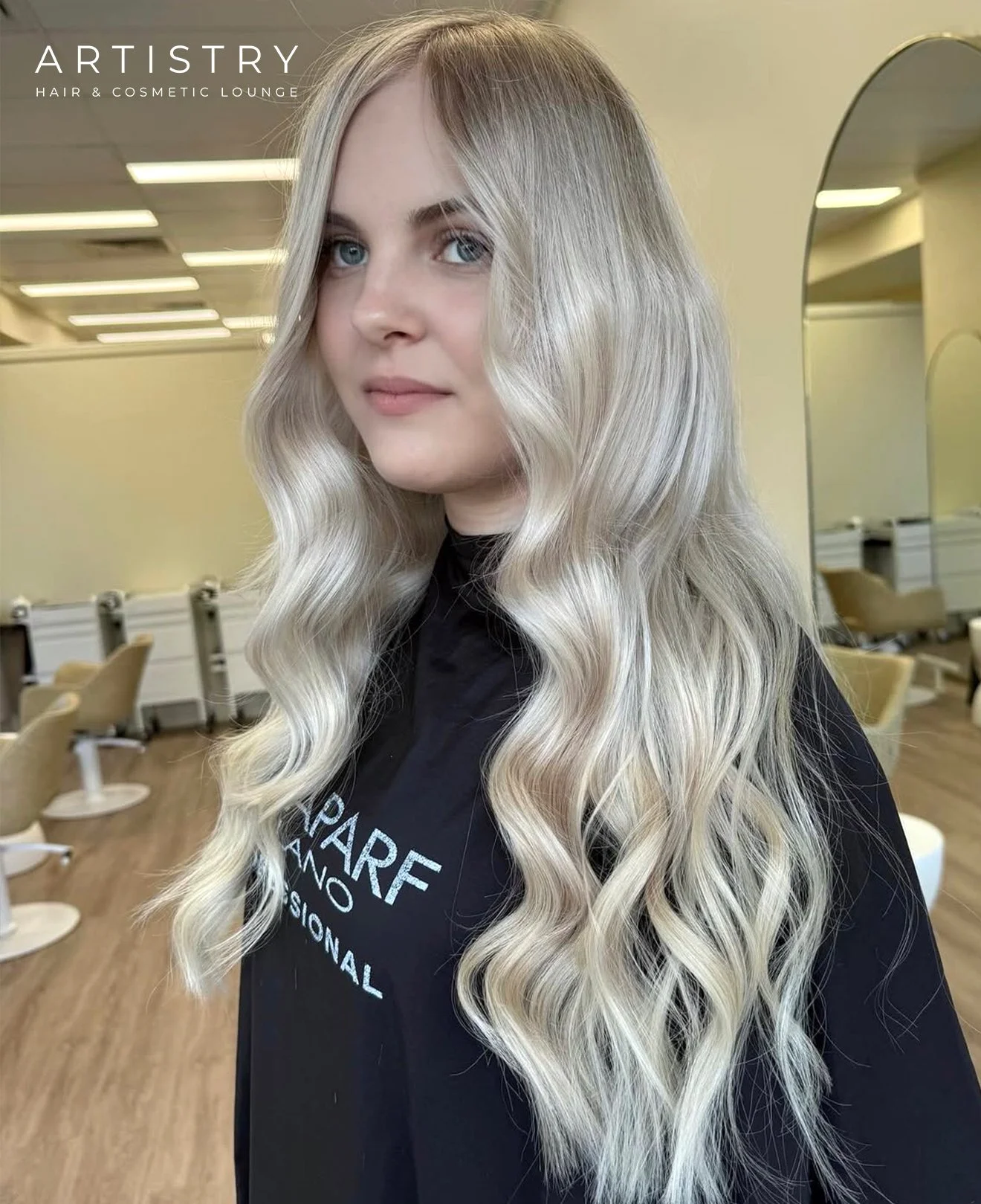 A woman with long, wavy blonde hair standing inside a salon with mirrors and chairs, wearing a black salon apron.