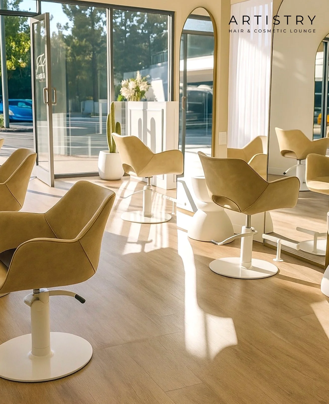Interior of a salon with beige chairs, large glass windows, and sunlight casting shadows on the wooden floor. There is a potted cactus plant and a small counter with flowers. The logo of Artistry Hair & Cosmetic Lounge is visible in the top right corner.