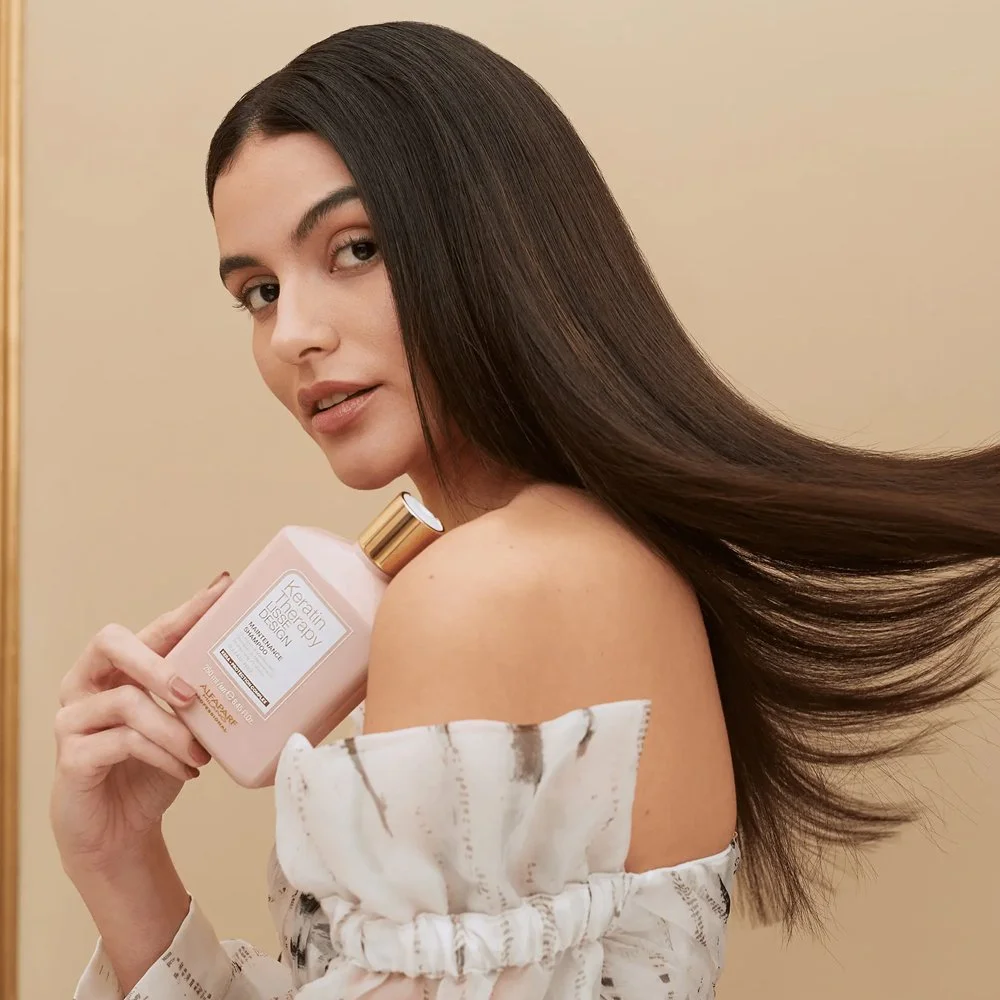 A woman with long, straight, dark brown hair holding a pink bottle of shampoo close to her shoulder, looking at the camera with a gentle expression.